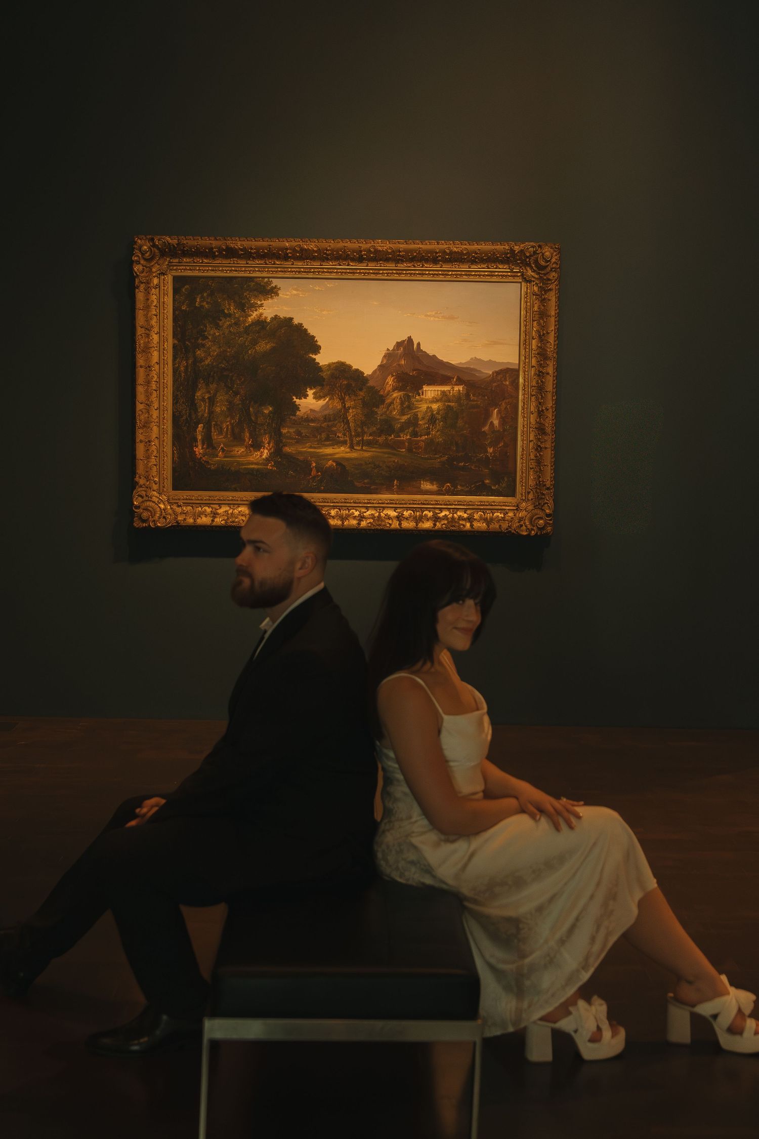 A couple sits back-to-back on a bench beneath a pastoral landscape painting in a museum.