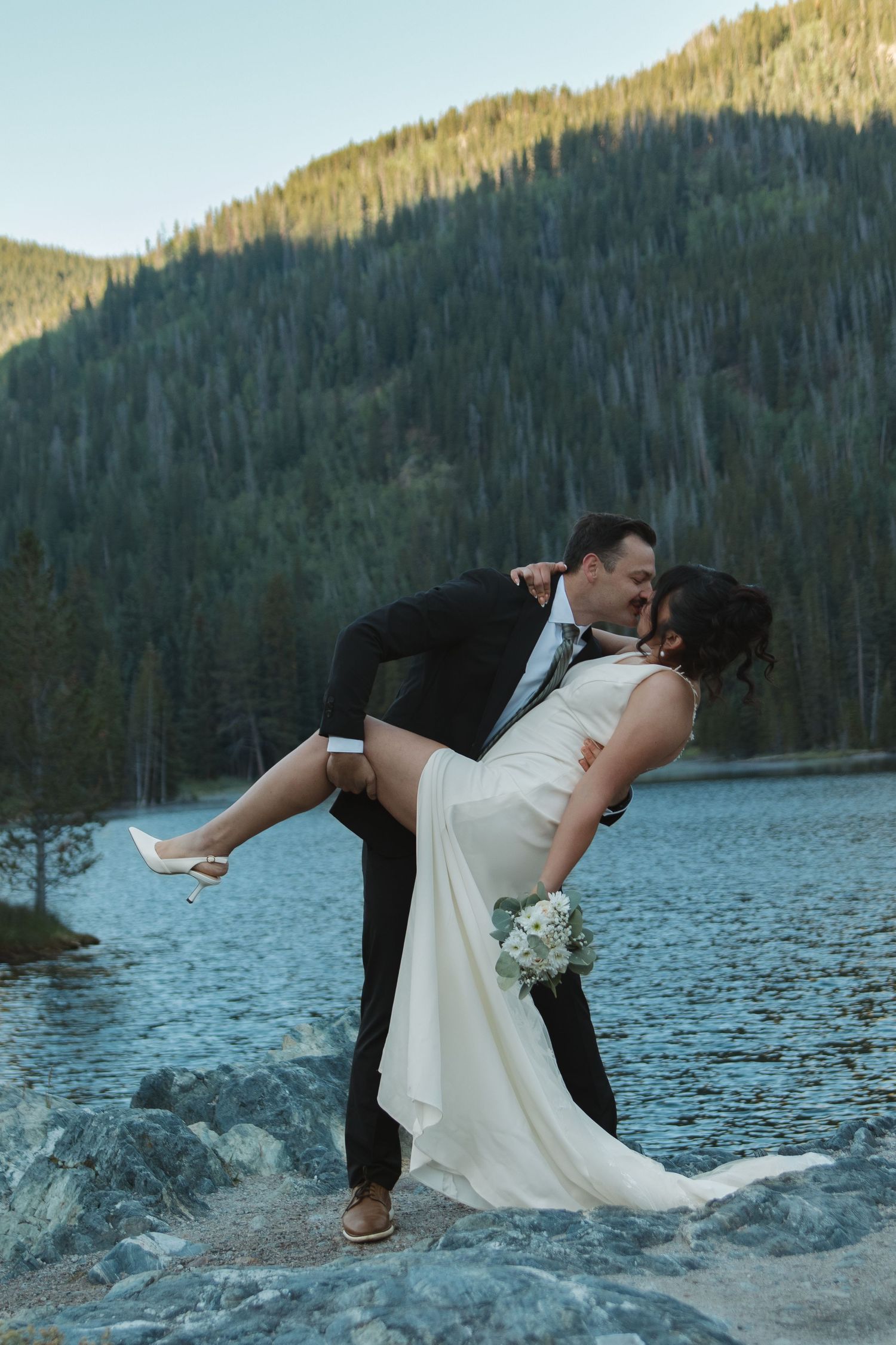 A wedding couple performs dramatic dip poses against a scenic mountain lake backdrop.