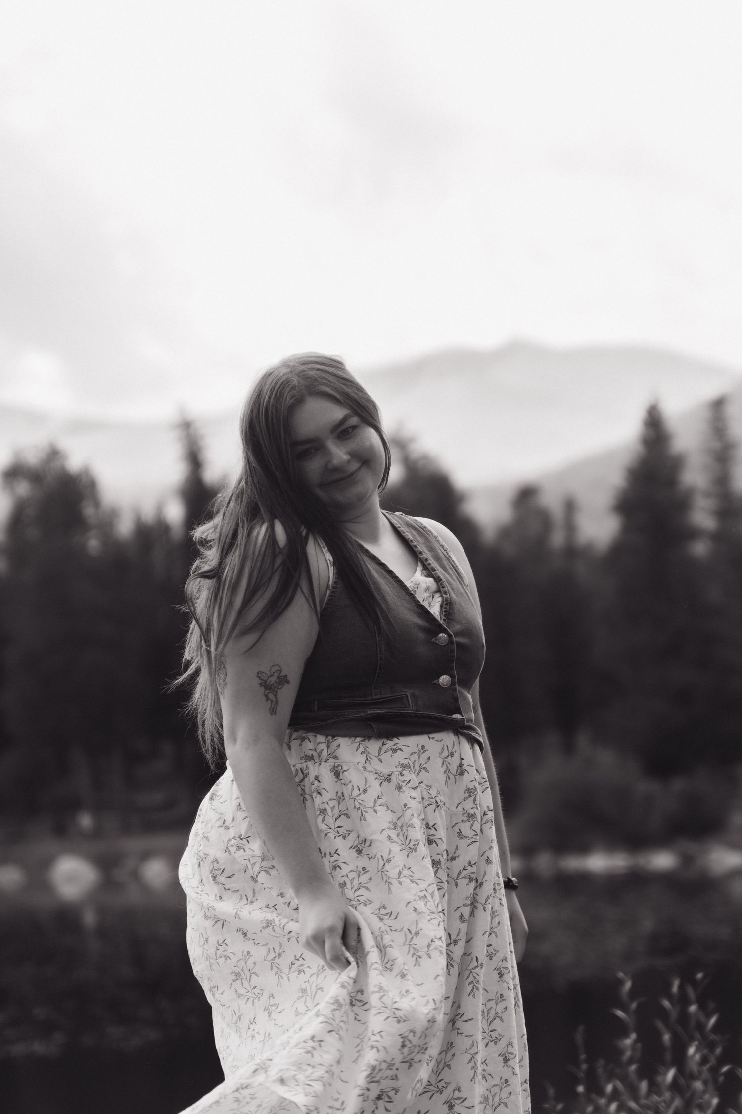 A person in a white lace dress stands in a field with evergreen trees and a lake in the background.