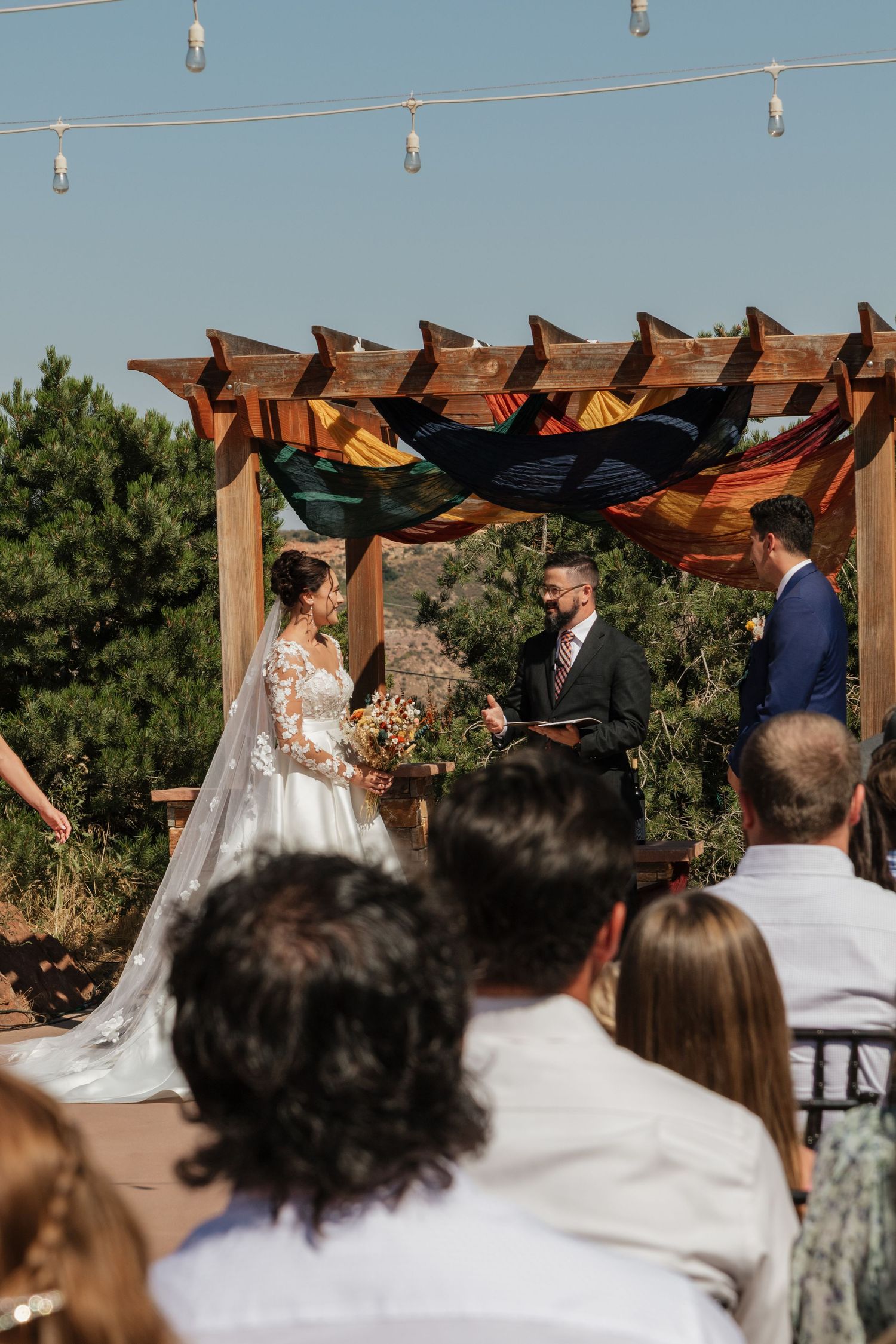 Outdoor wedding ceremony under wooden pergola with guests seated and mountains in background.
