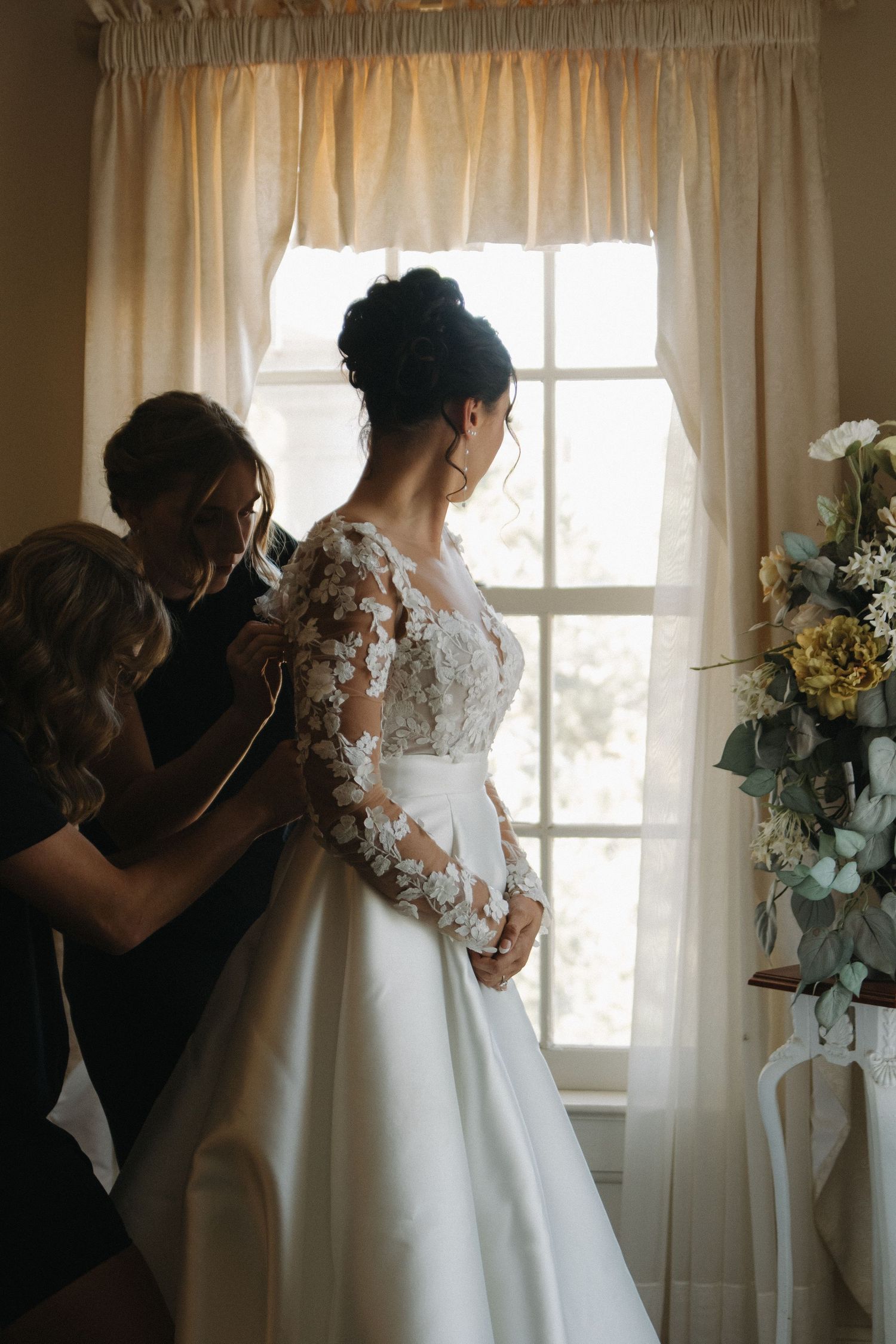 Silhouette of wedding dress preparations near a sunlit window with flowing curtains.