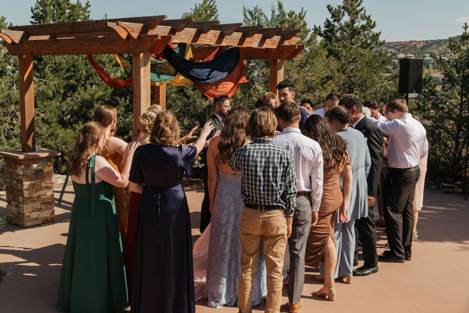 Scenic outdoor wedding gathering under wooden pergola with guests in summer attire.