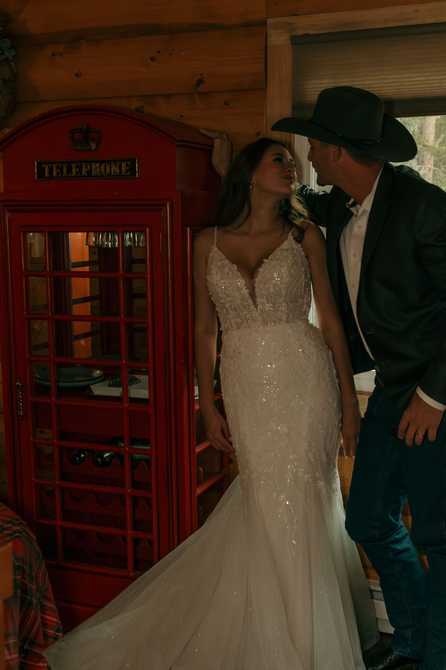 A couple poses near a vintage red telephone booth in a rustic setting.