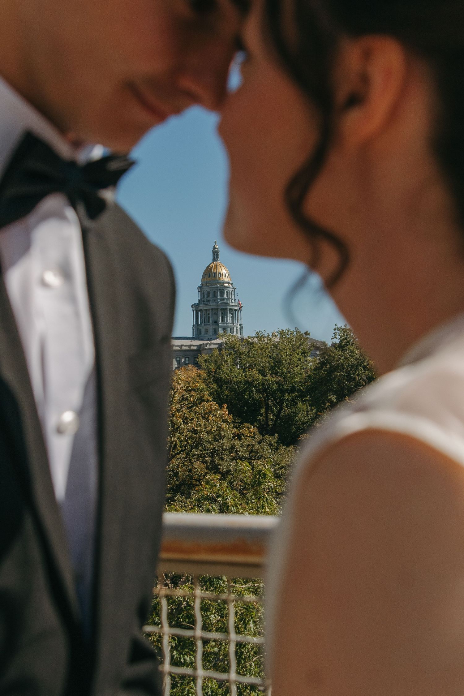 Wedding couple silhouetted against bright blue sky with trees and building in background.