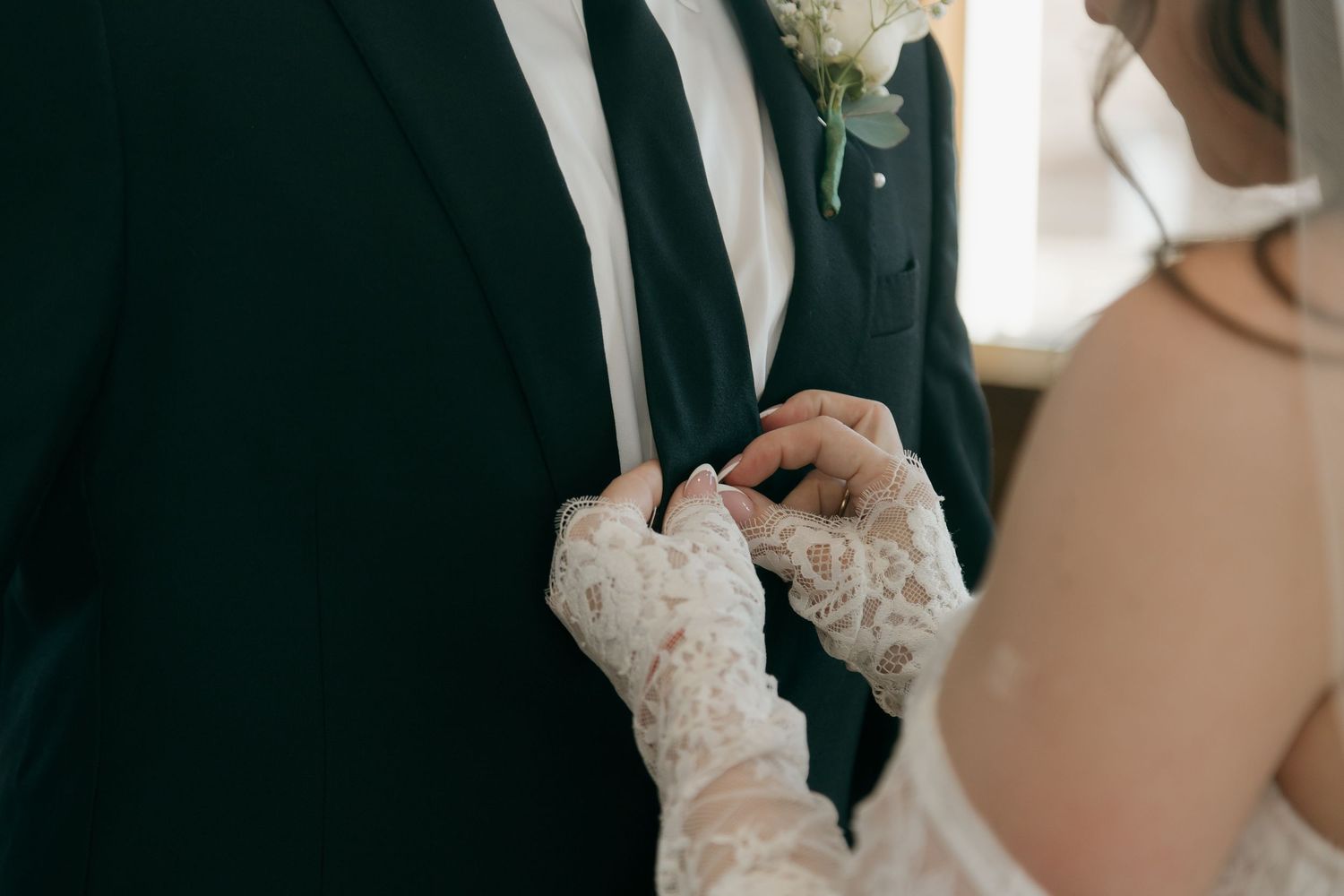 Bride in lace gloves adjusting grooms black tuxedo lapel before wedding ceremony.