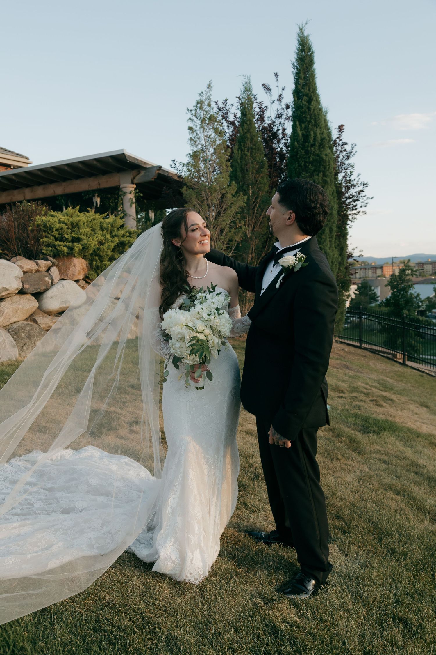 Bride in elegant white mermaid gown with flowing veil and groom in black tuxedo sharing a romantic moment outdoors.
