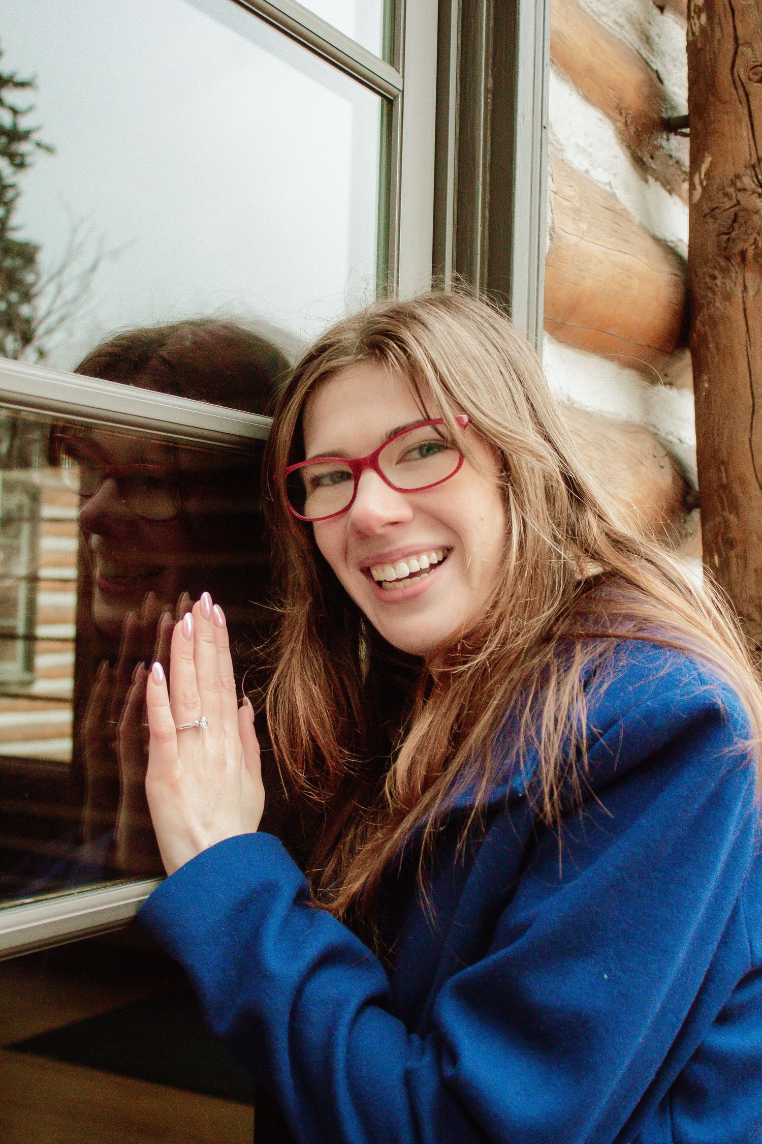 A person in a blue coat and red glasses looks through a cabin window with joy.