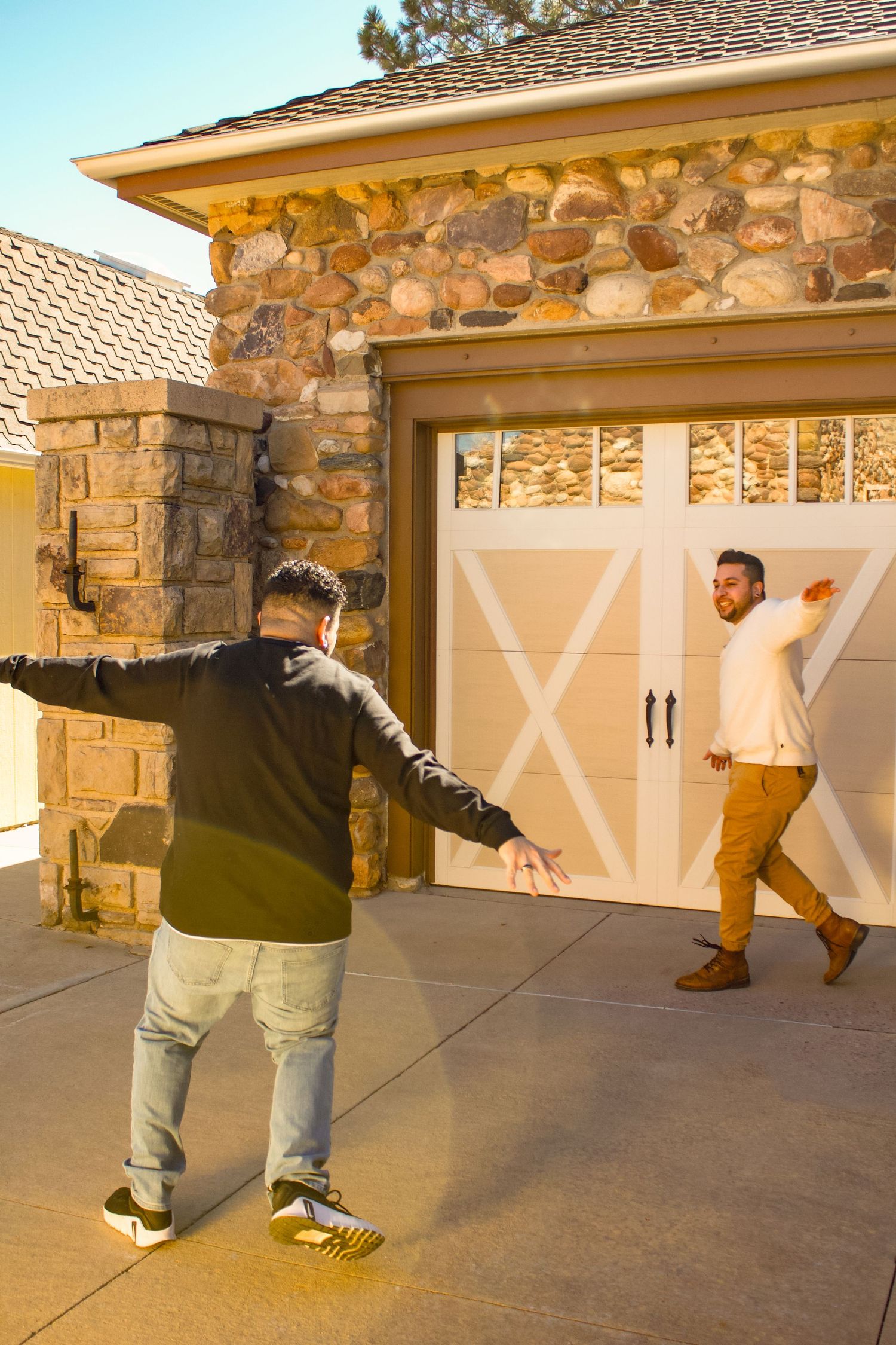 Two people pose playfully in front of a stone facade house with white barn-style garage doors at sunset.