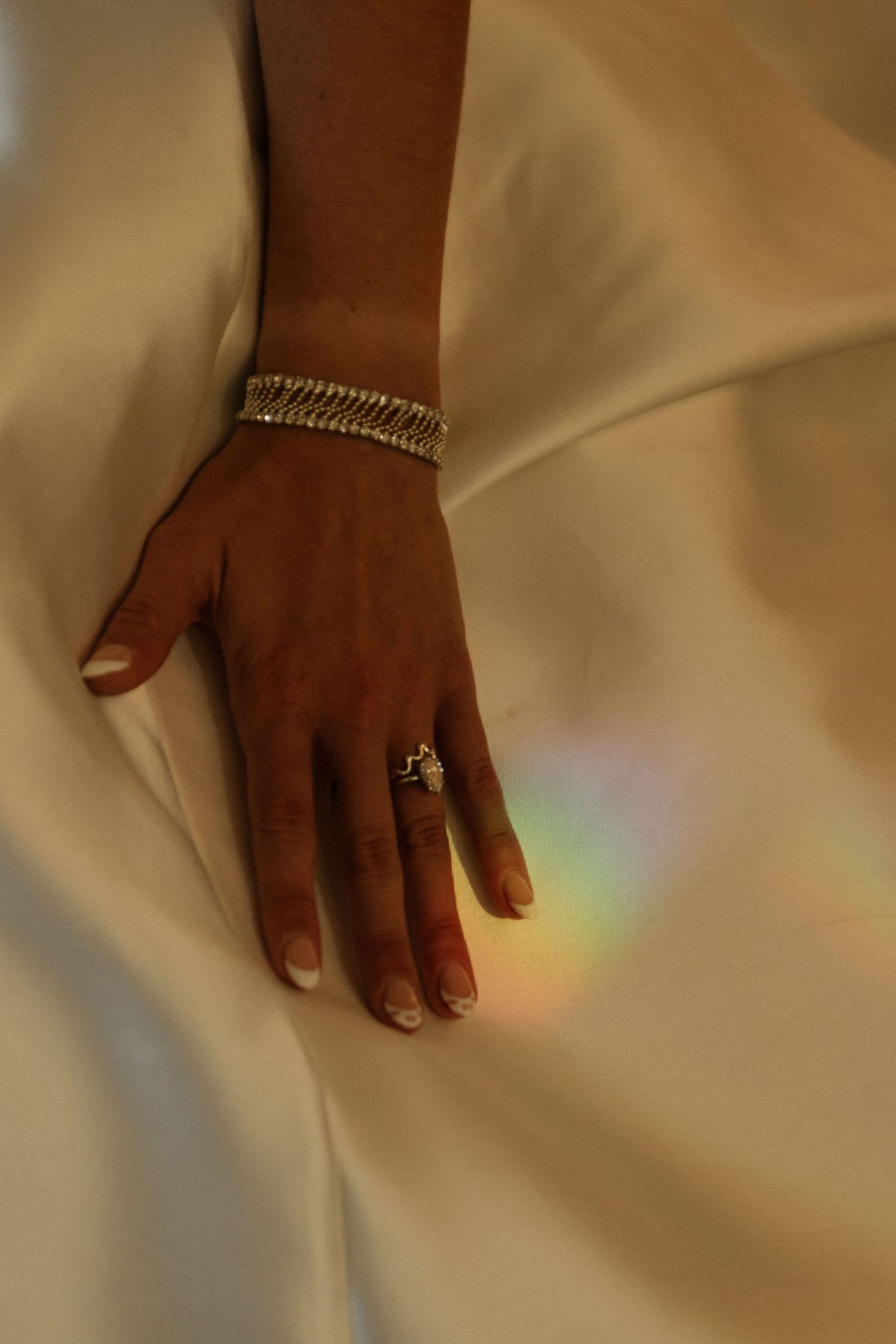 A hand wearing a delicate silver ring and bracelet rests on a cream-colored satin fabric with a subtle rainbow reflection.