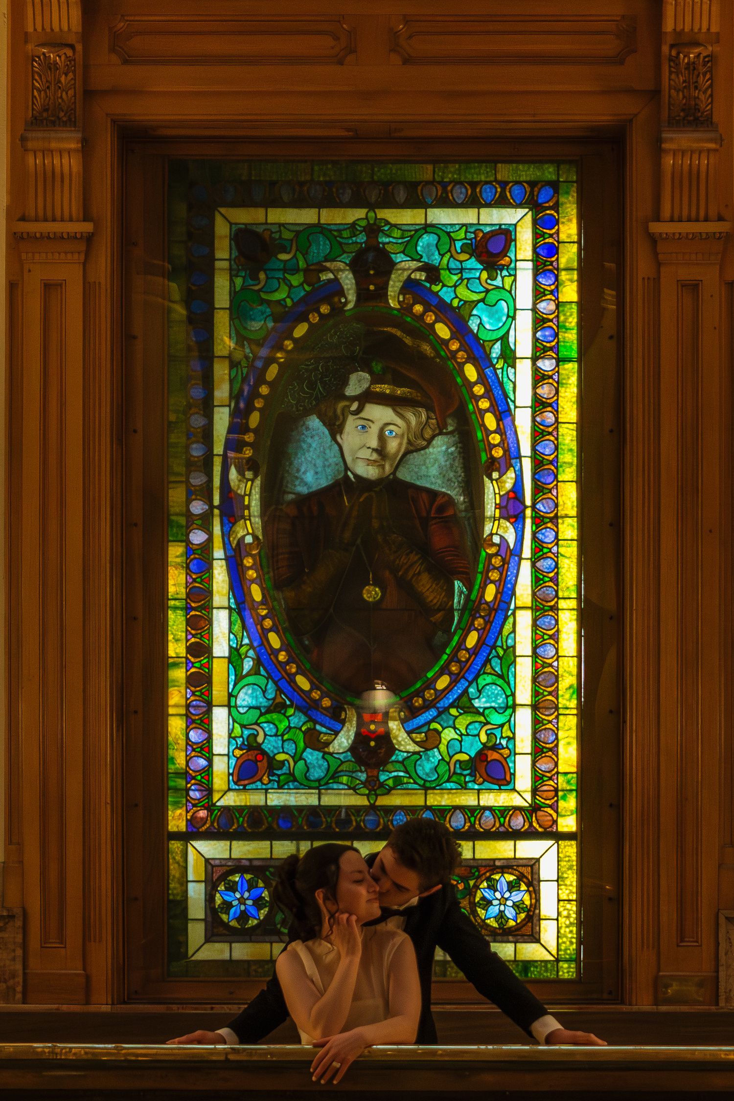 A couple poses in front of colorful stained glass windows in an ornate interior setting.