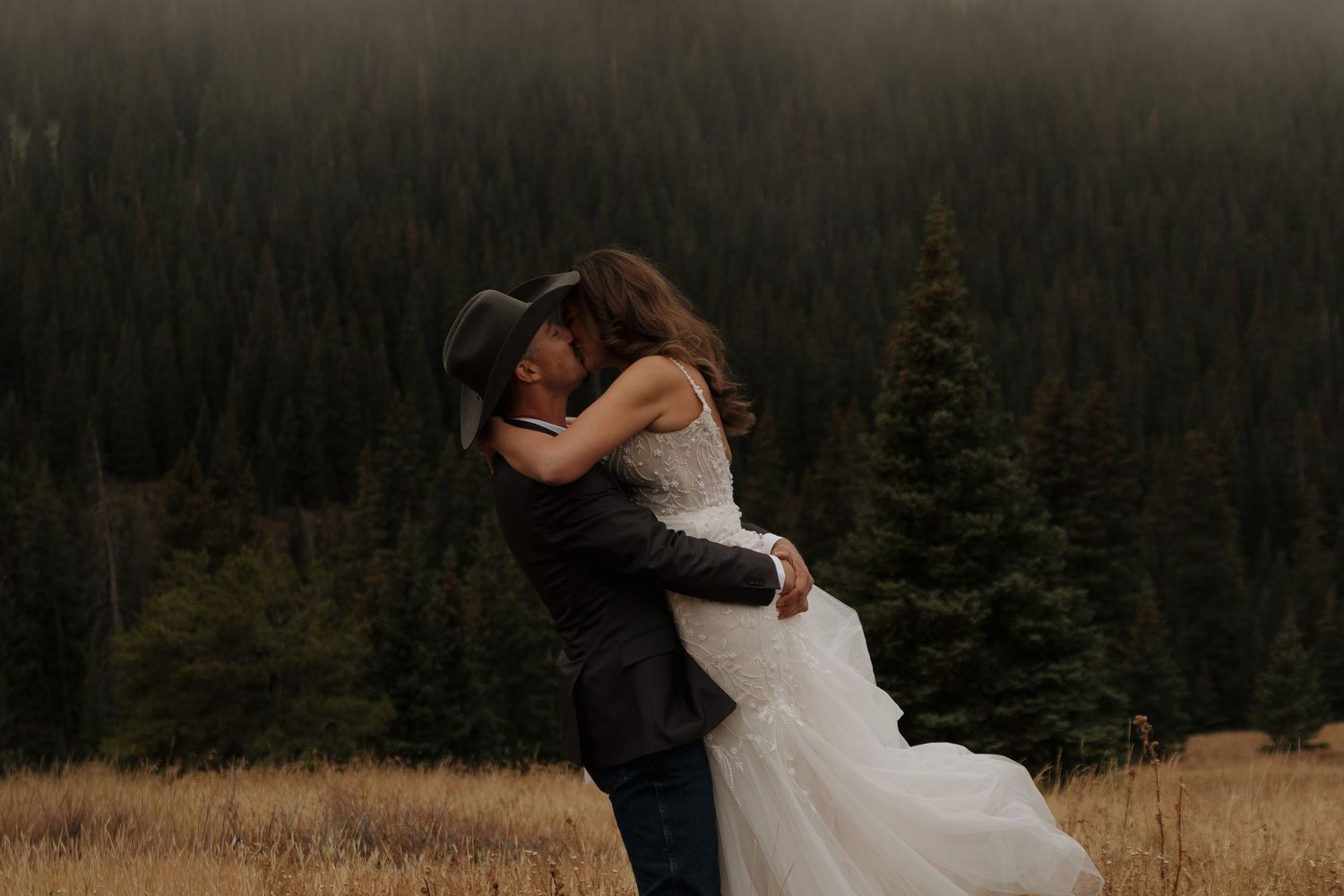 A romantic embrace on a mountain meadow with evergreen forests and misty peaks in the background.