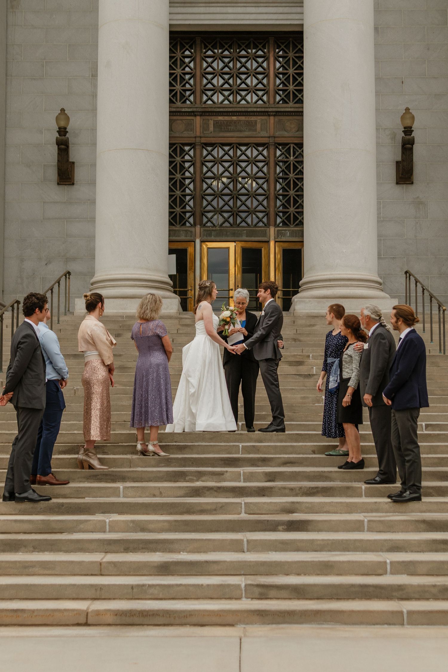 Wedding party gathers on the steps of a grand building with white columns and ornate architectural details.