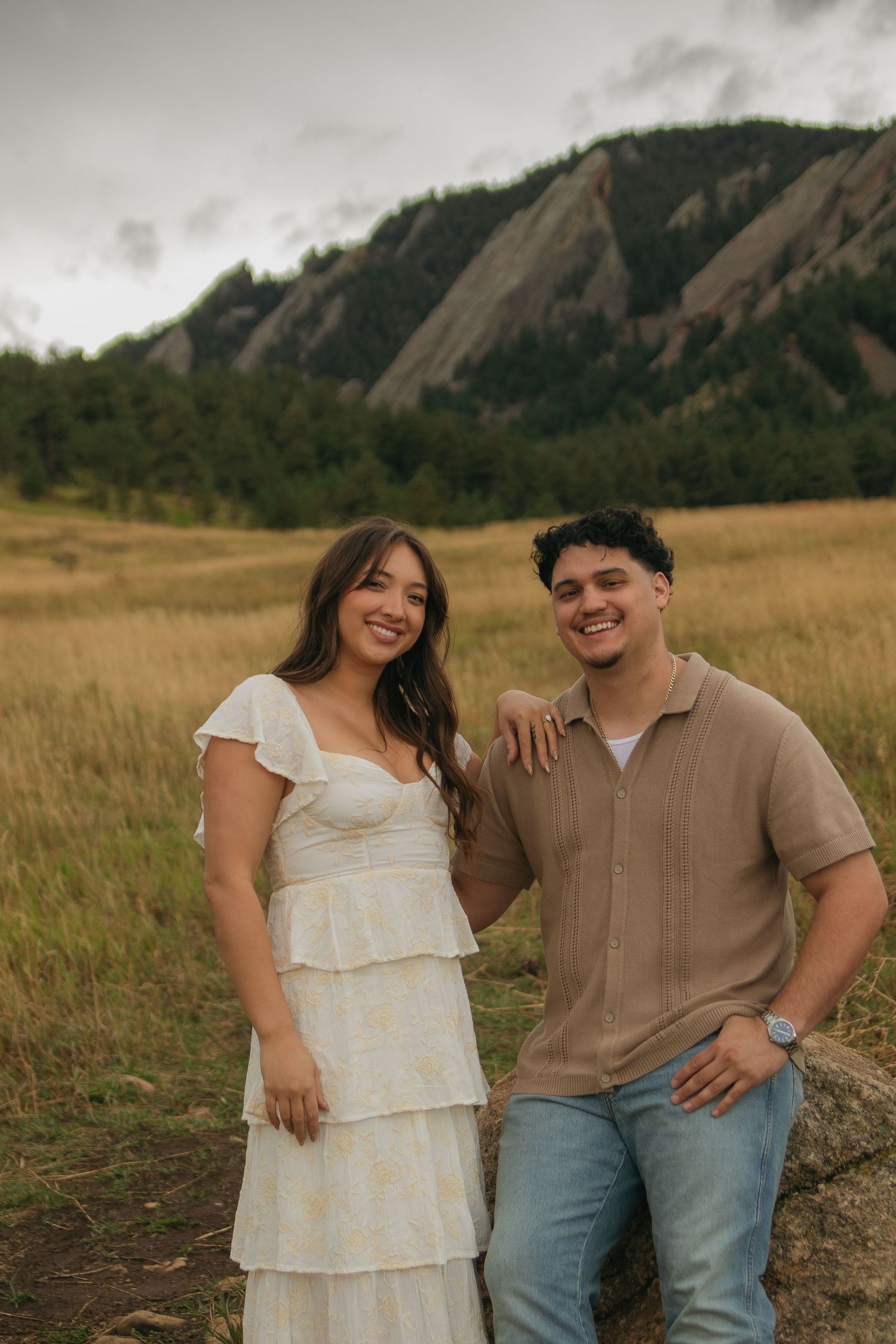 Smiling couple leaning on rock in grassy field with rocky mountain flatirons in background.