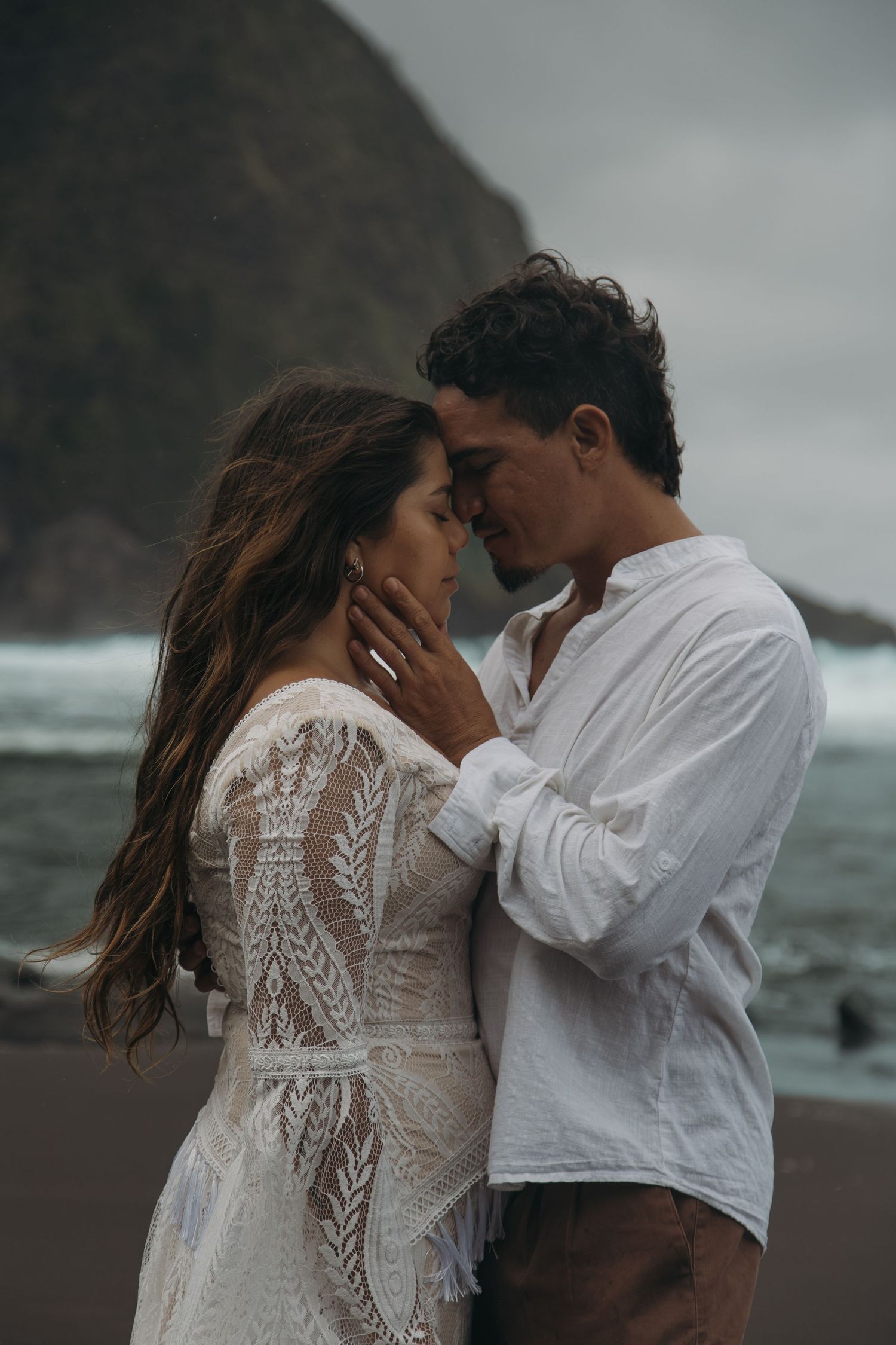 Intimate couple embrace on black sand beach, bride in lace boho gown with rocky cliffs behind.