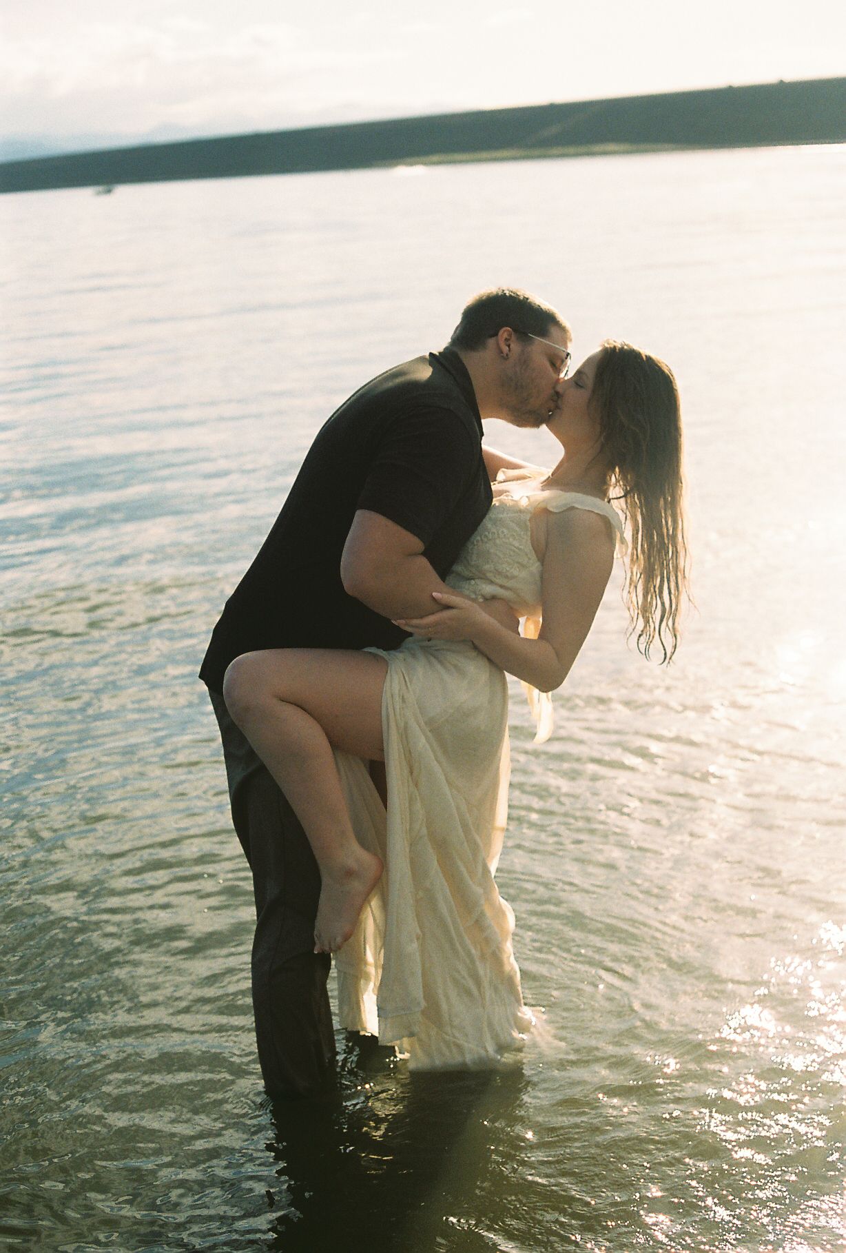 Couple sharing passionate kiss in lake water at sunset, man dipping woman in wet white dress.