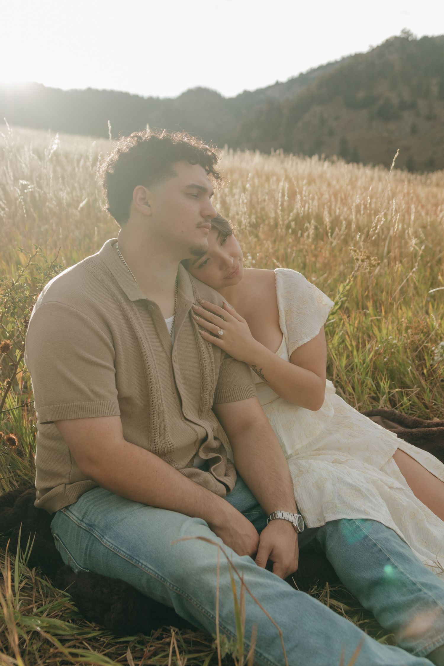 A couple sits together in a sunlit golden field, the woman resting her head on the mans shoulder.