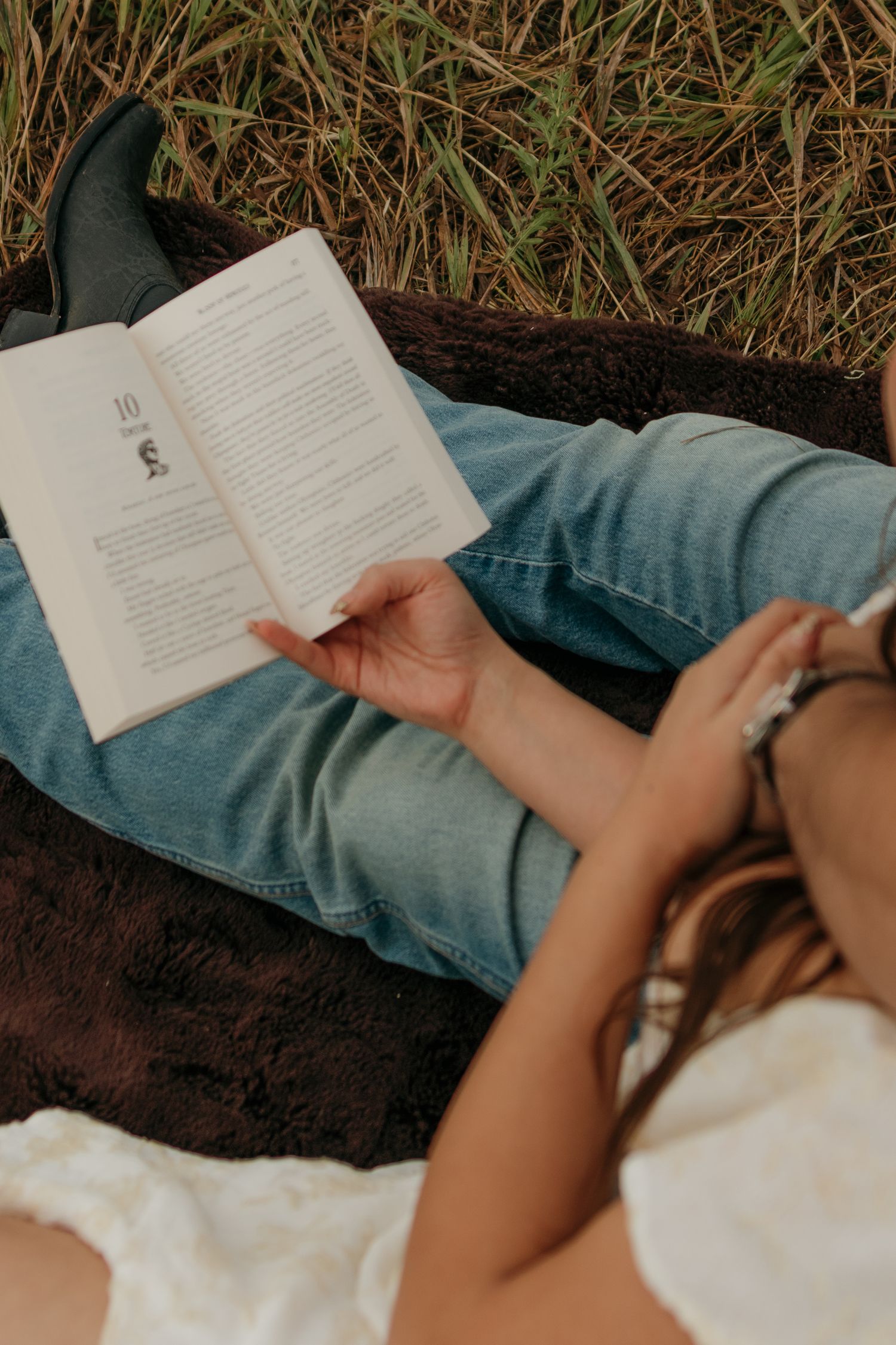 Person reading a book while relaxing outdoors on a blanket in a grassy field wearing jeans and boots.
