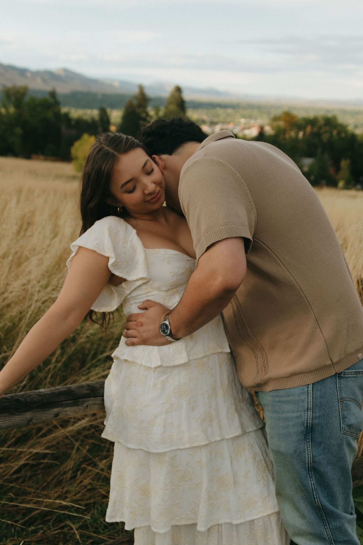 Man tenderly embraces woman in white tiered dress by wooden fence in scenic meadow.