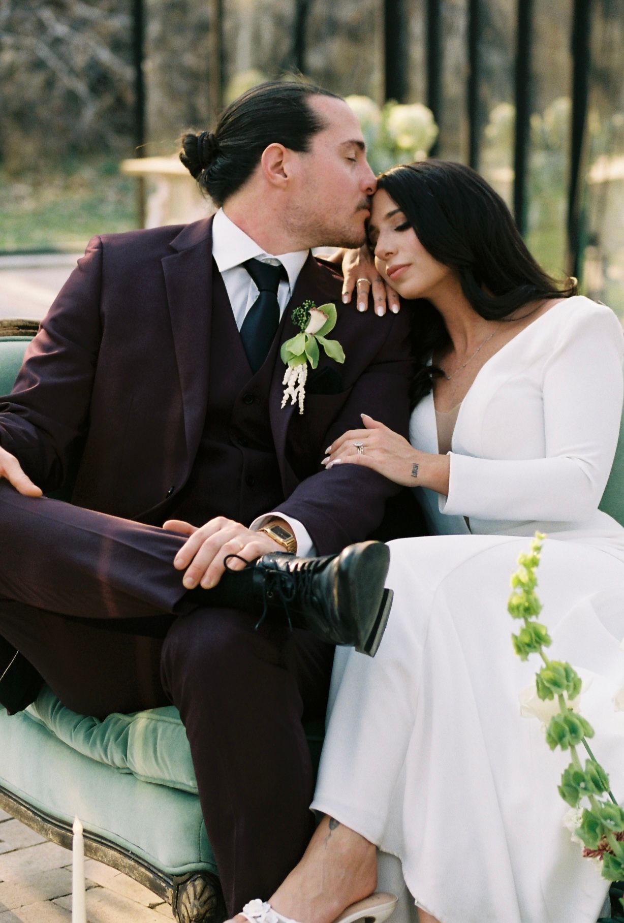 Groom in burgundy suit kisses bride's temple as they sit together on a vintage sofa at outdoor wedding.