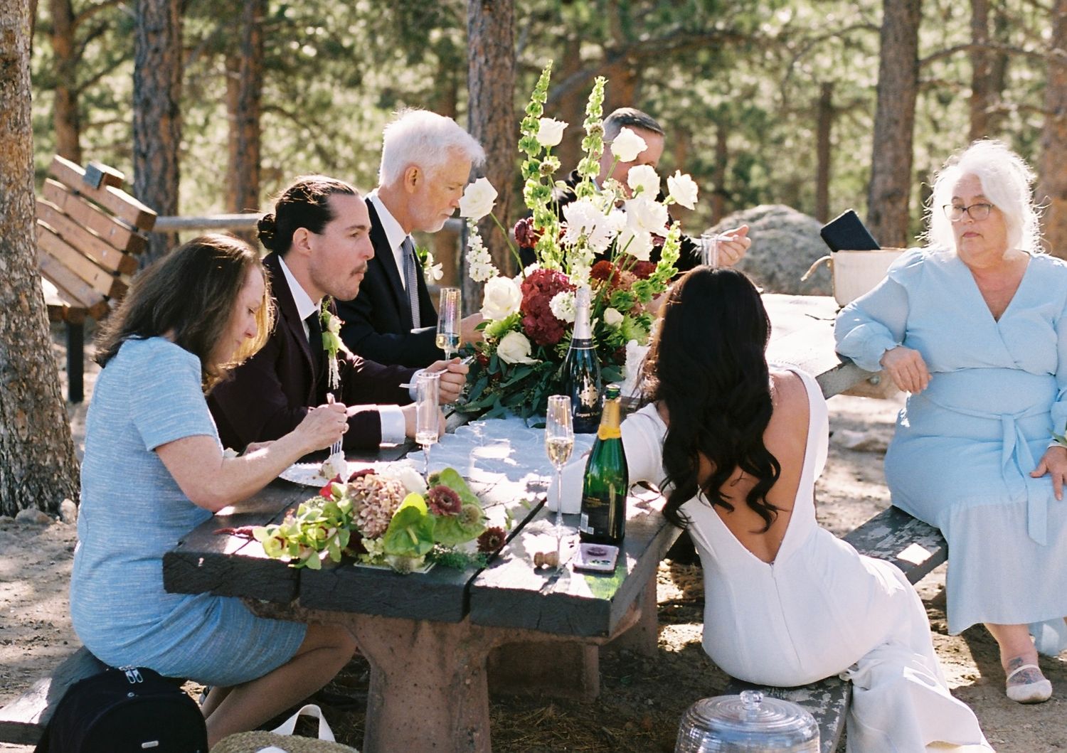 Intimate elopement reception with family gathered at wooden picnic table in pine forest.