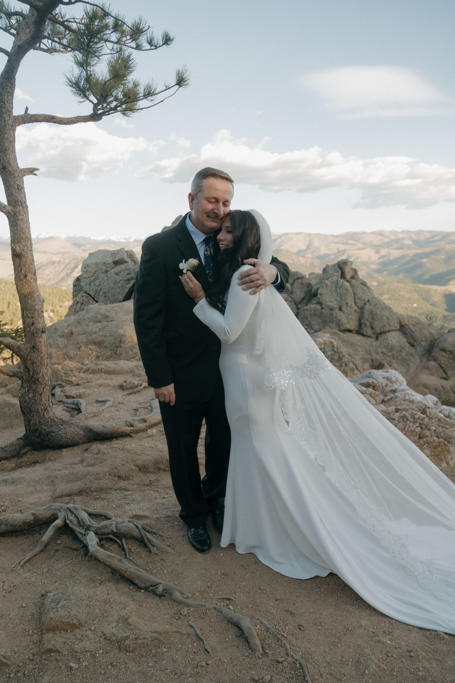 Bride and groom sharing a tender embrace on a scenic mountain summit with pine tree.