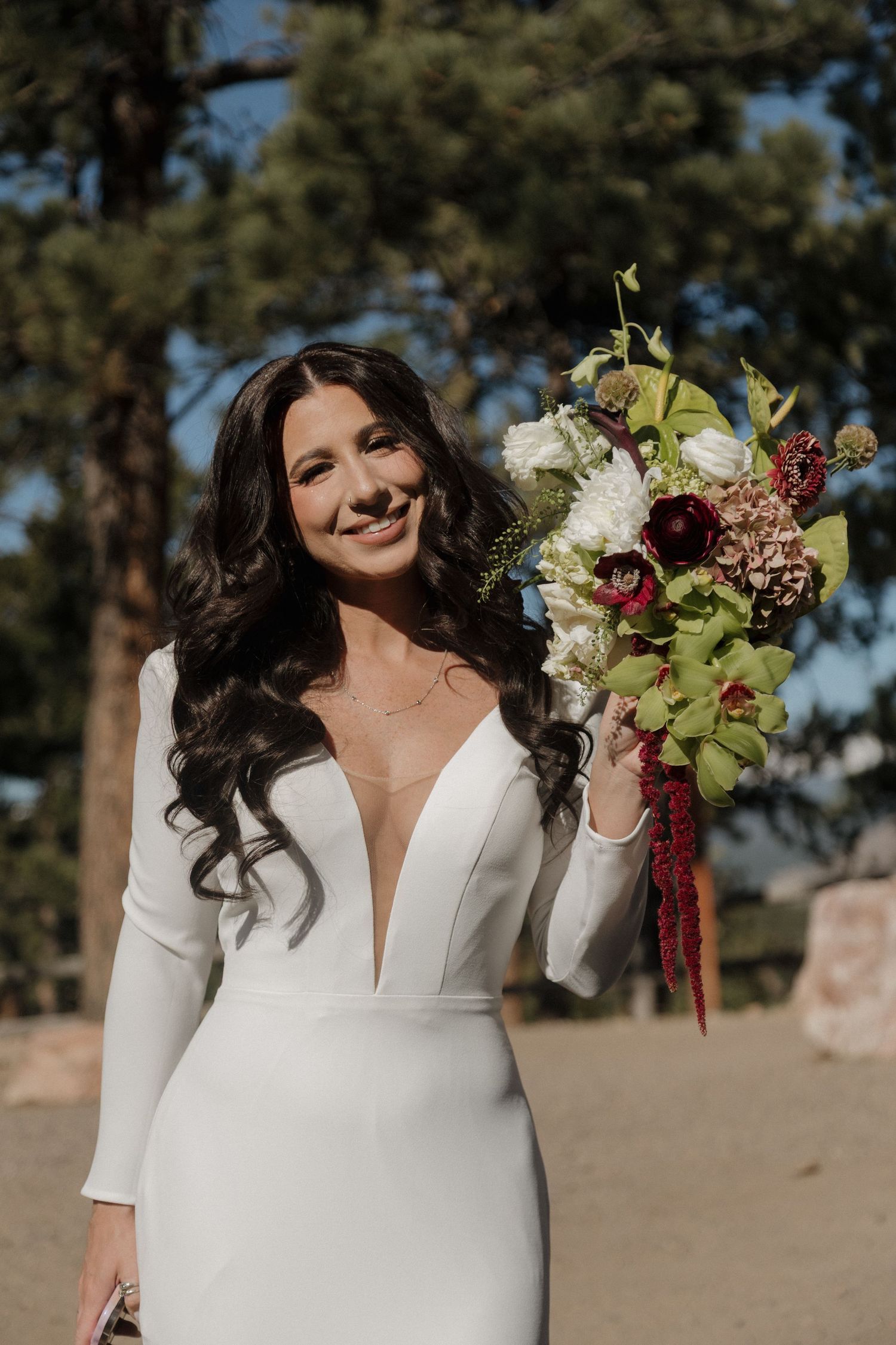 Smiling bride in white deep-V gown holding lush bouquet of green, white, and burgundy flowers outdoors.