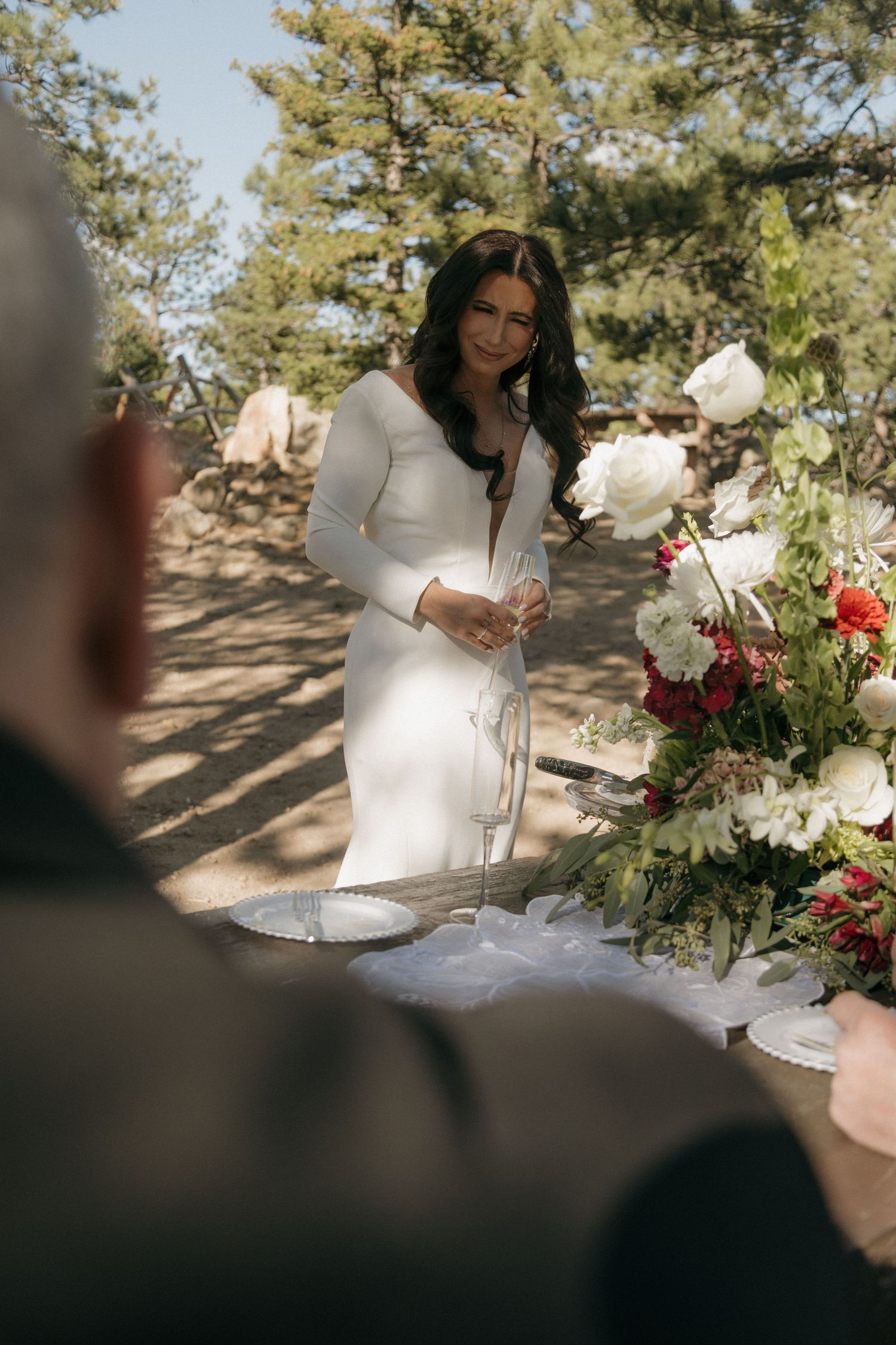 Bride in white long-sleeve gown stands at floral ceremony table during outdoor wedding.