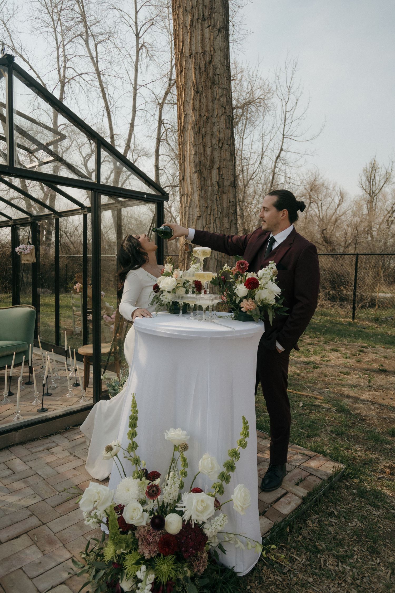 Bride and groom pour champagne tower outdoors near a glass greenhouse wedding venue.