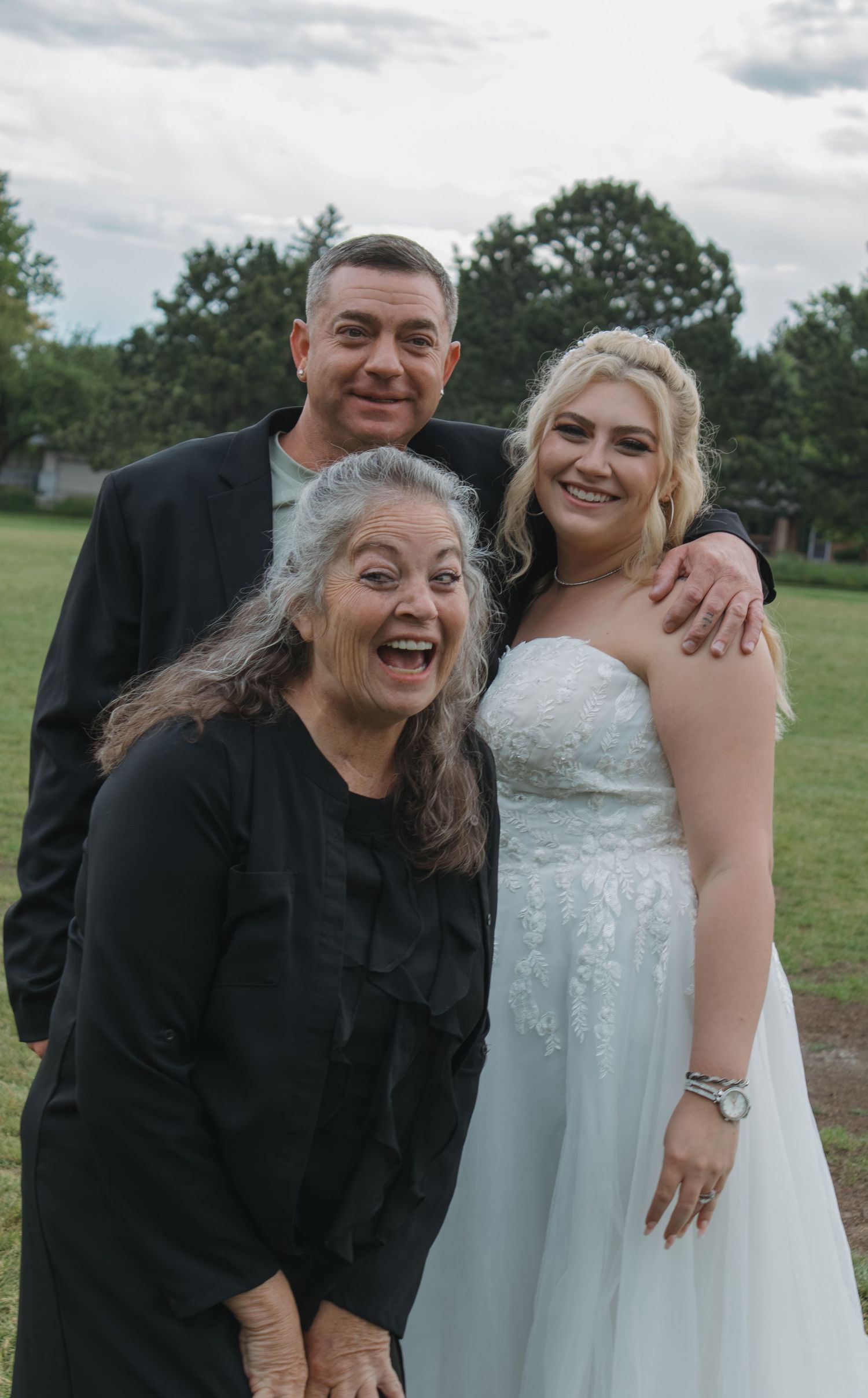Three people share a joyful moment together while standing on grass with trees in the background.