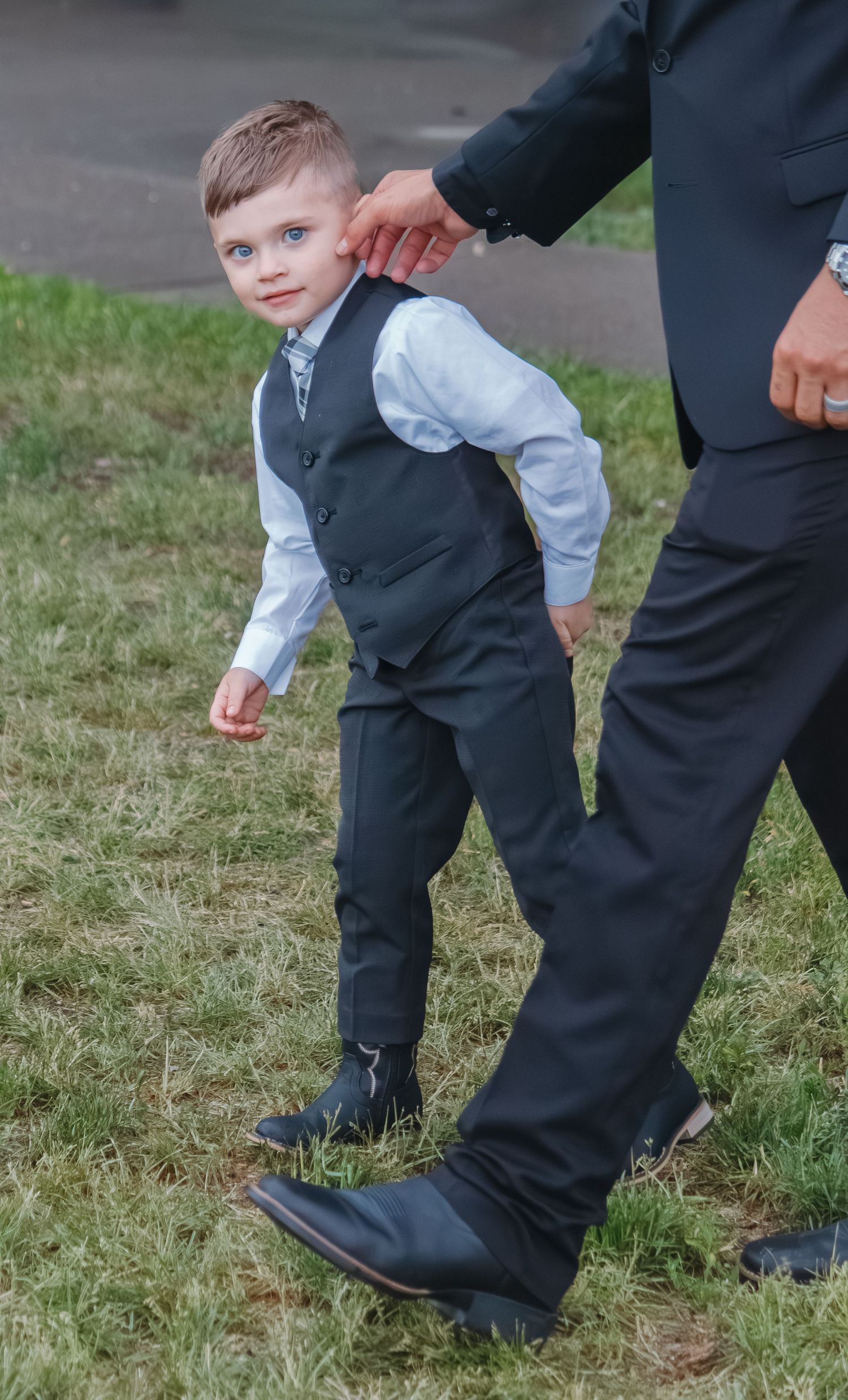 A young ring bearer in formal attire standing next to an adult at an outdoor event.