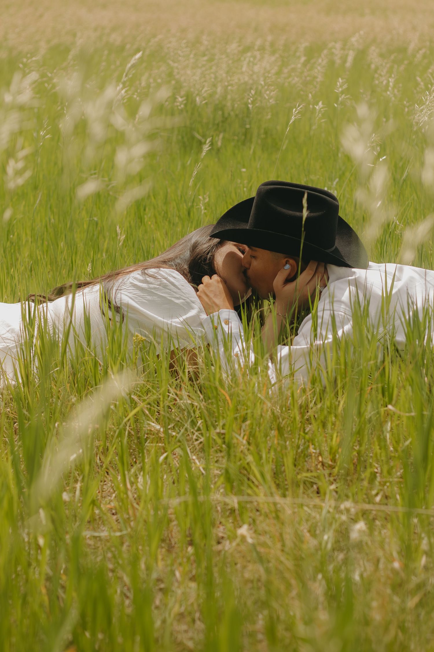 A couple shares an intimate embrace while lying in tall wild grass under natural lighting.