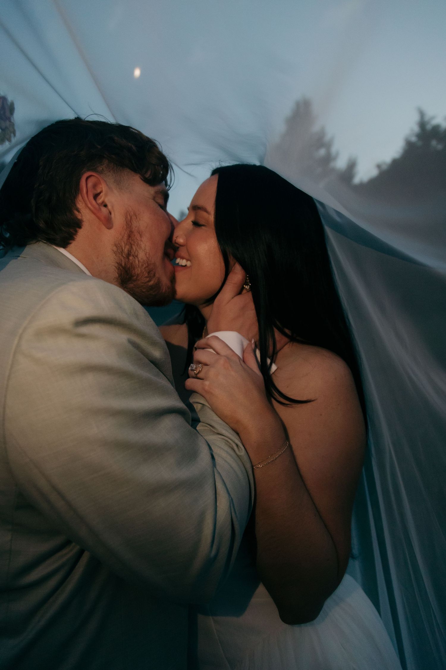 Groom kissing smiling bride under flowing veil at dusk with moonlit forest backdrop.