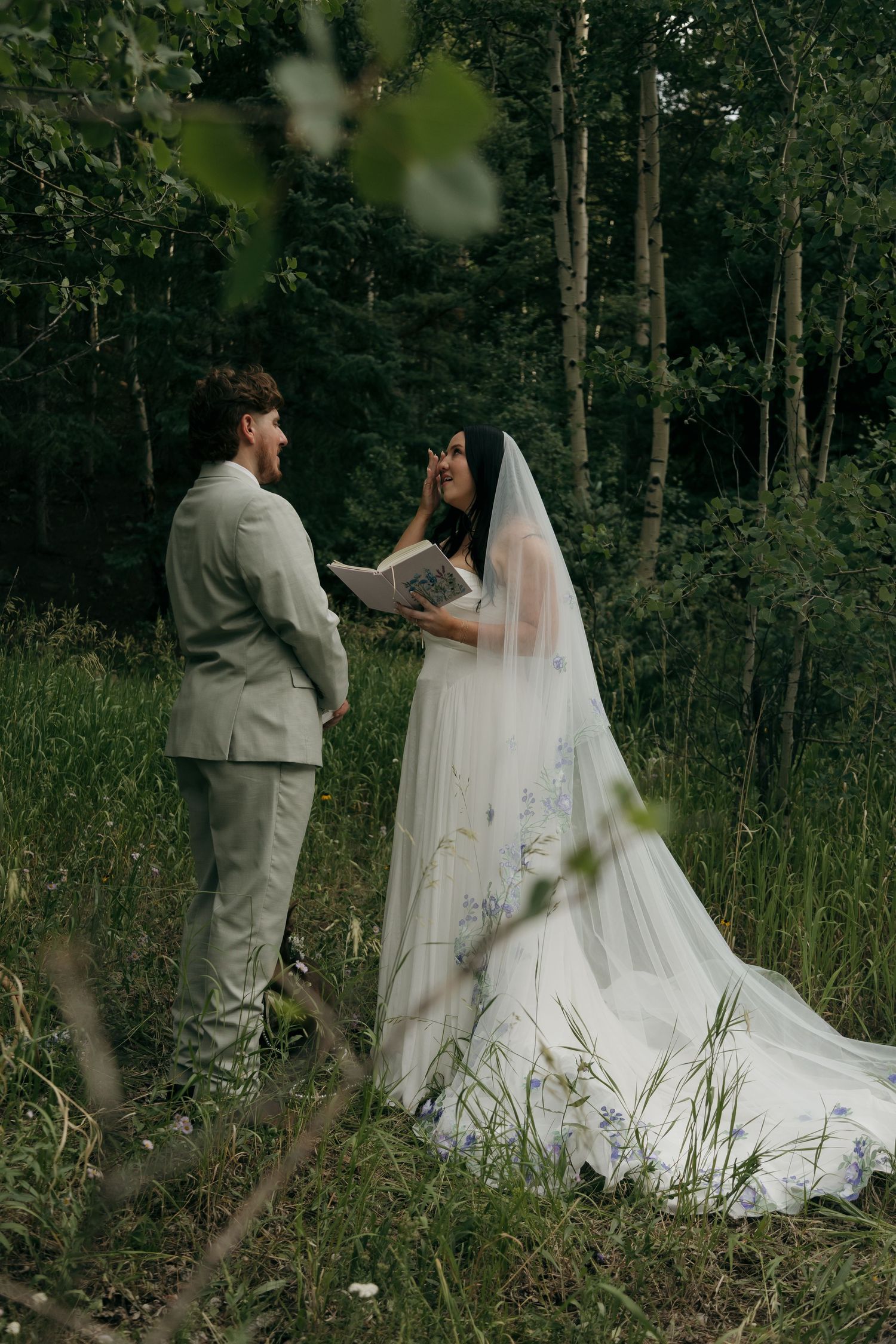 Bride reads vows to groom in lush forest clearing, wearing white gown with blue floral details and cathedral veil.