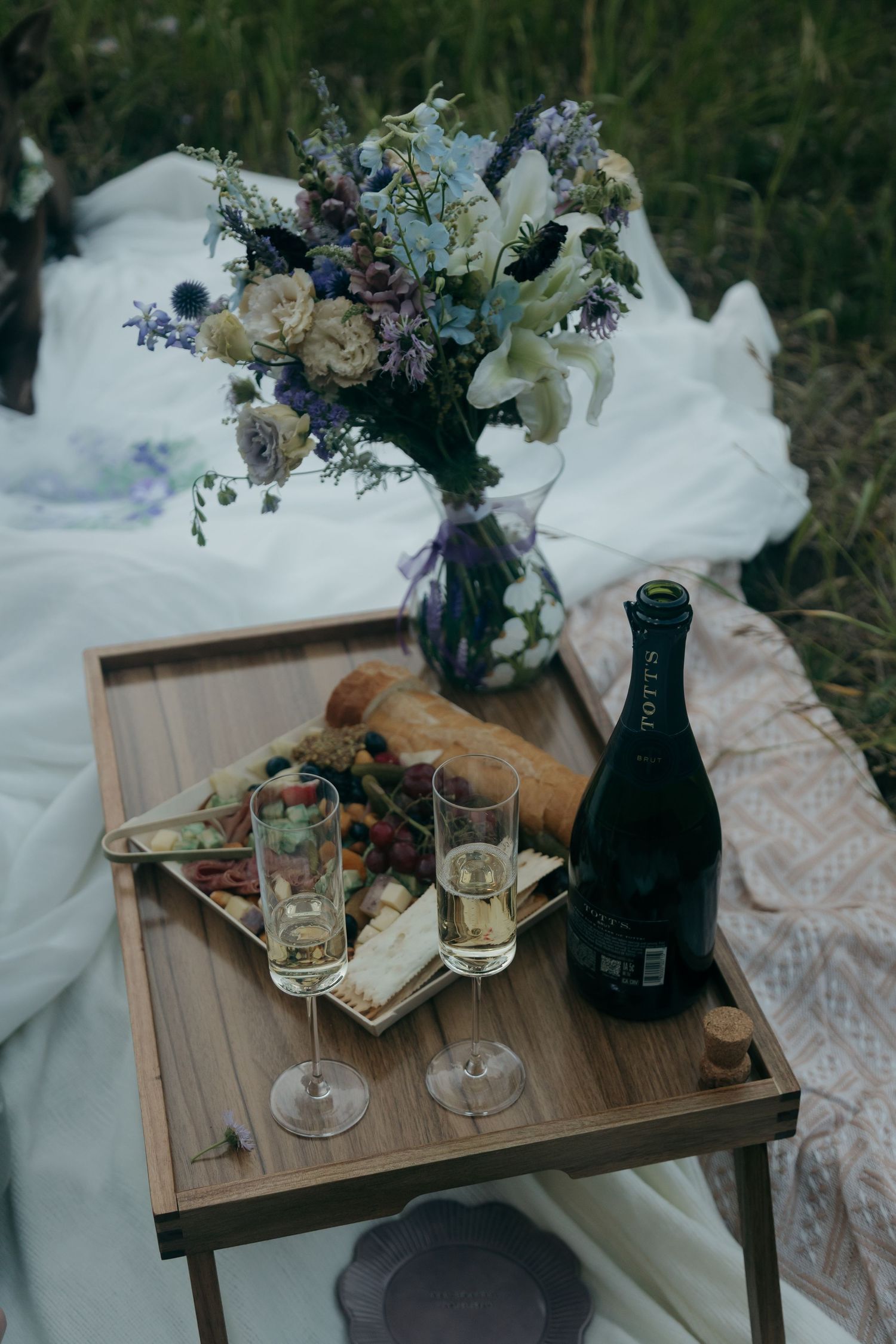 Elegant outdoor wedding picnic with champagne, charcuterie, baguette, and wildflower bouquet.