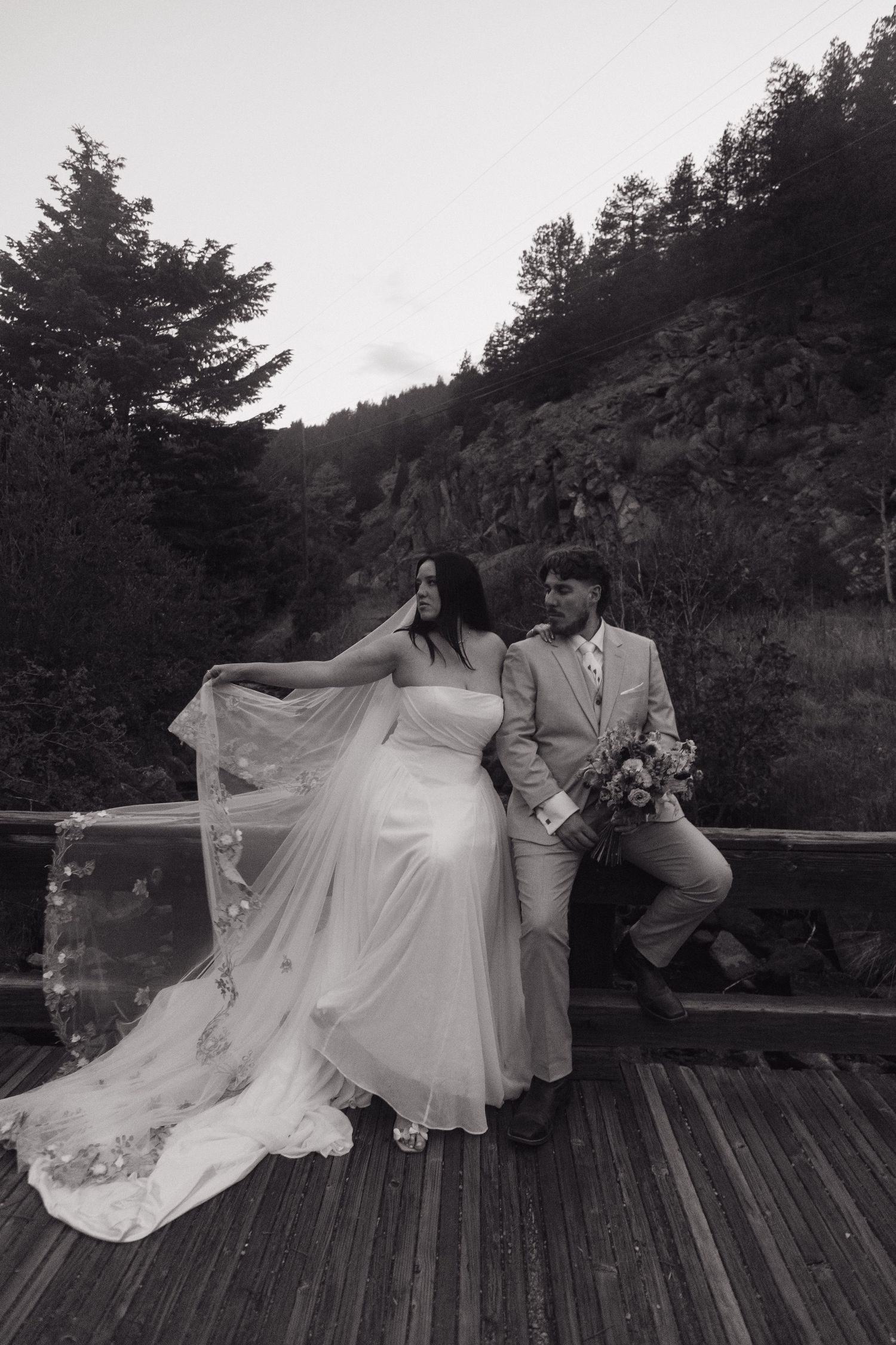 Black and white portrait of bride and groom on rustic wooden bridge with mountain forest backdrop at twilight.