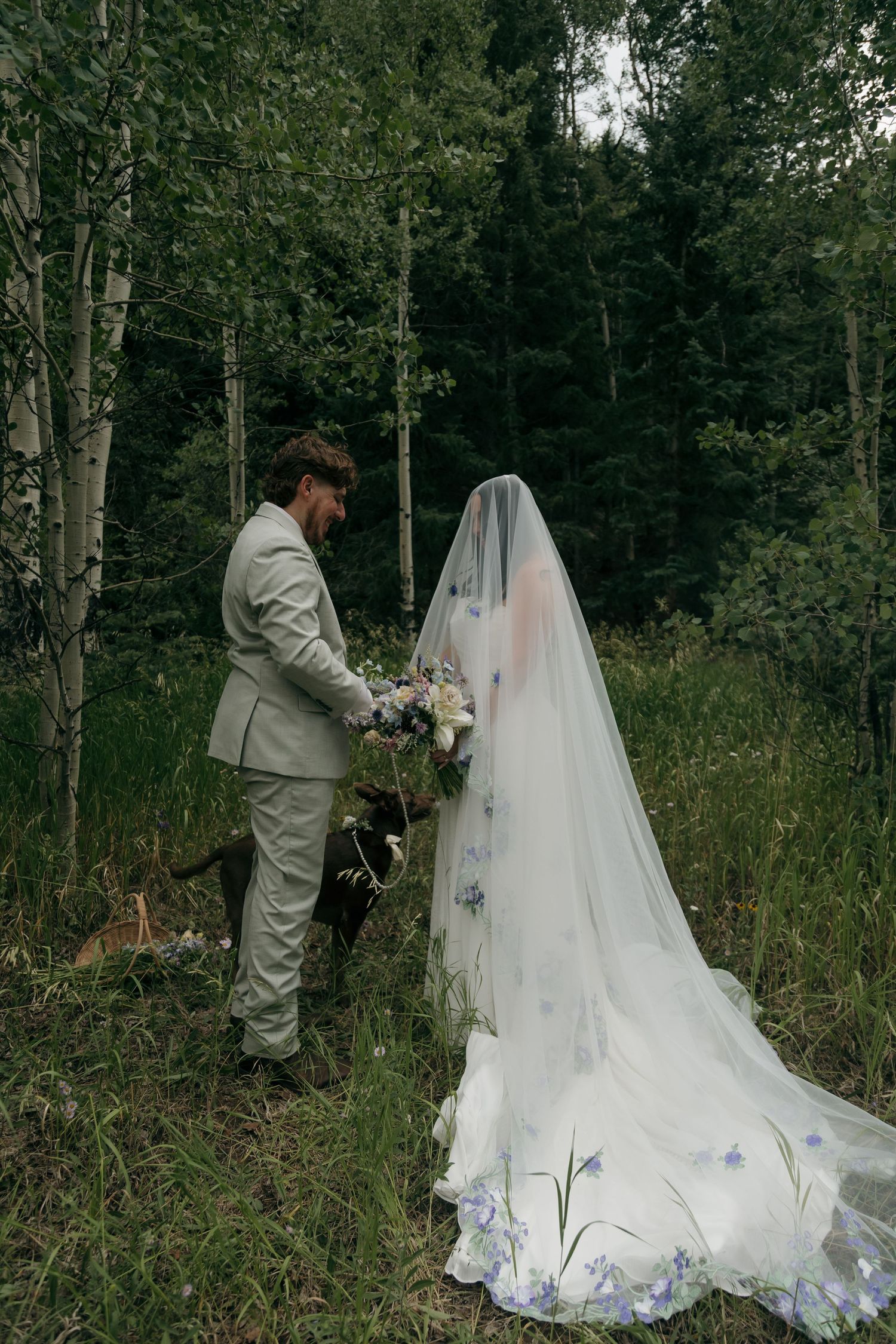 Bride in floral veil and groom in gray suit stand with dog in lush forest wedding photo.