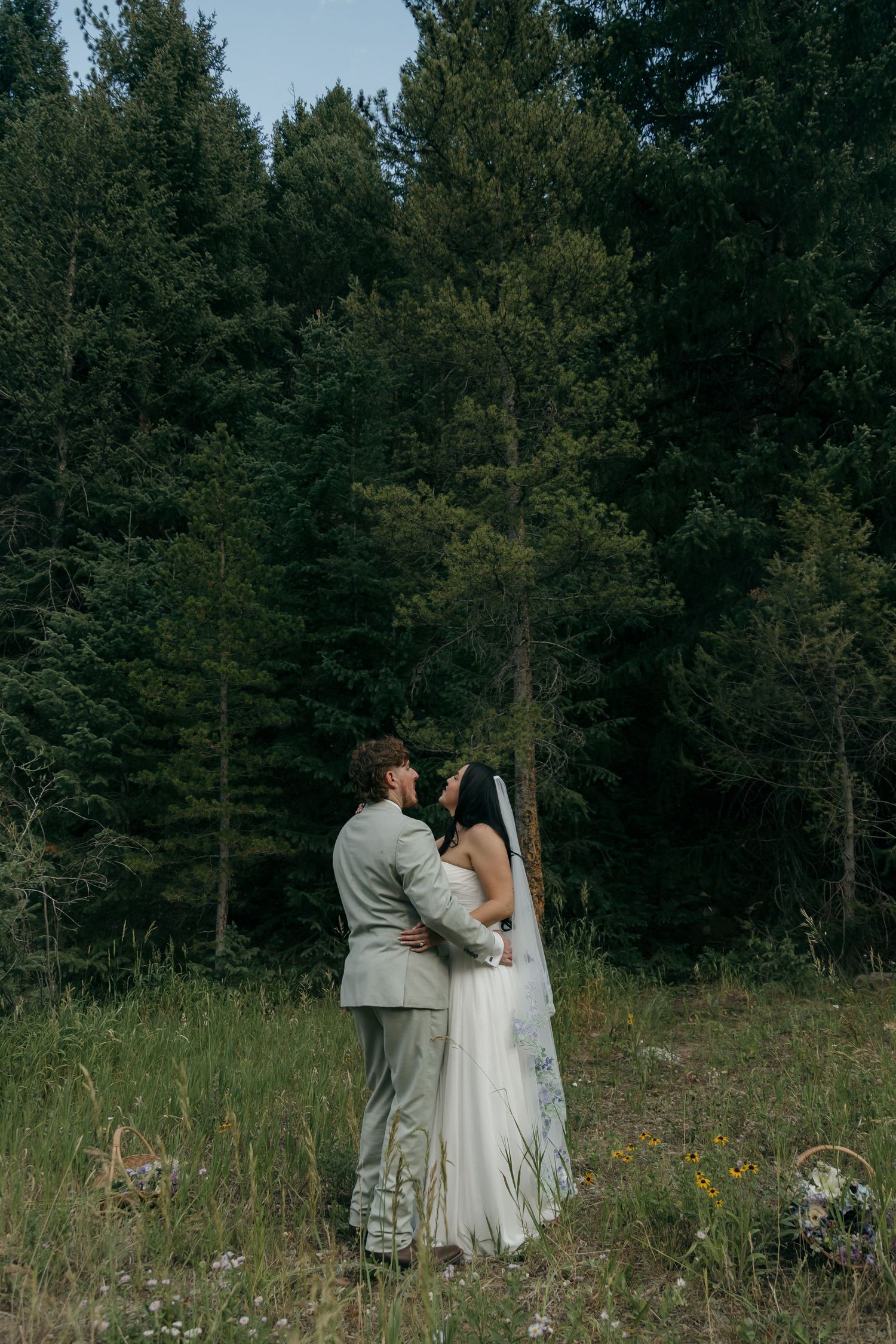 Bride and groom embrace in a wildflower meadow surrounded by tall evergreen trees during outdoor forest wedding.