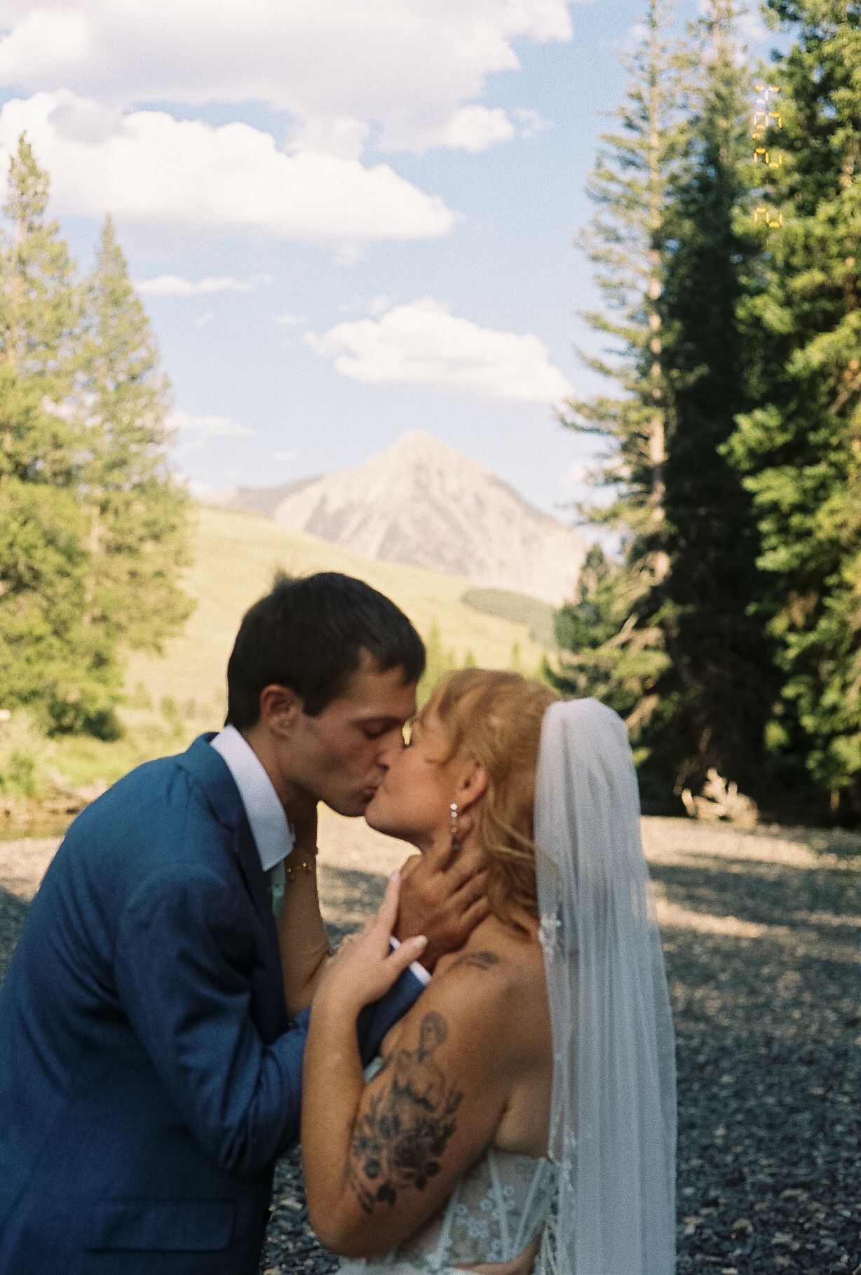 Bride and groom kissing outdoors with tall pine trees and mountain peak under blue sky.