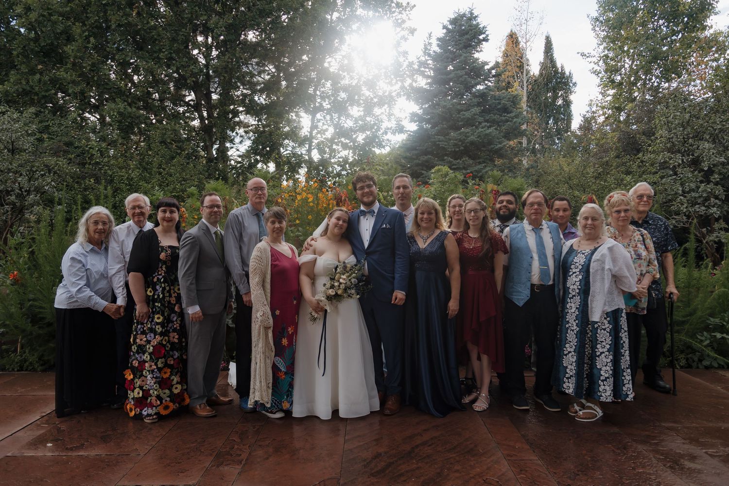 Wedding couple poses with family and friends in a lush garden setting, bride holding bouquet in white gown.