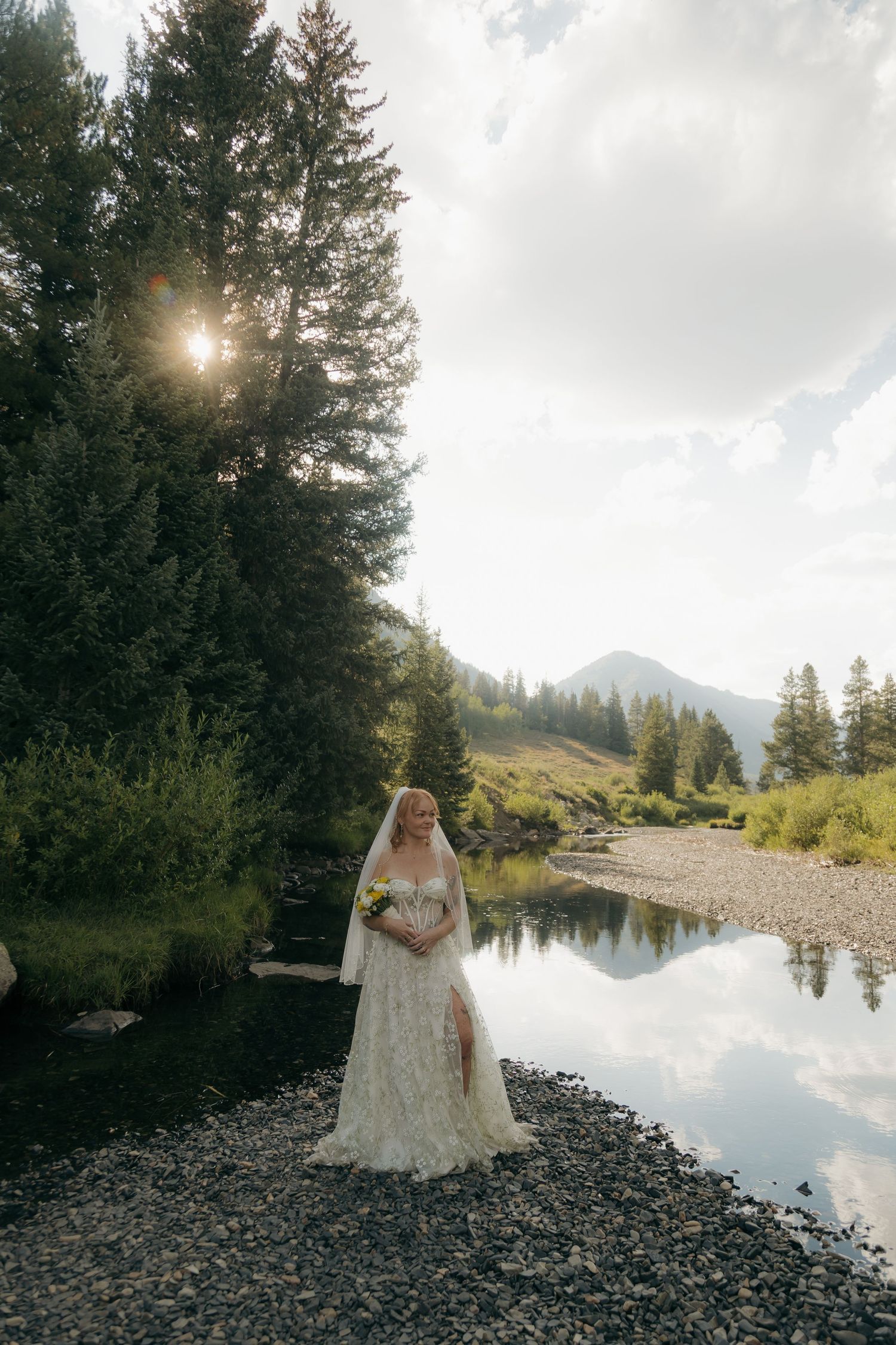 Bride in lace wedding dress and veil stands by mountain stream with sun rays through pine trees.