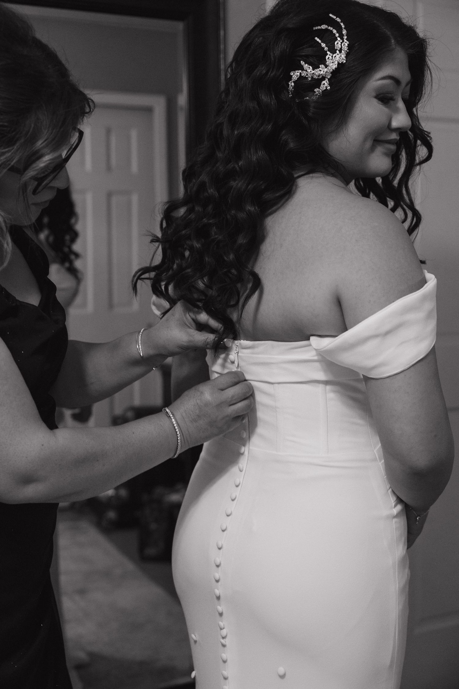 Black and white photo of bride in off-shoulder gown having buttons fastened by helper before wedding.