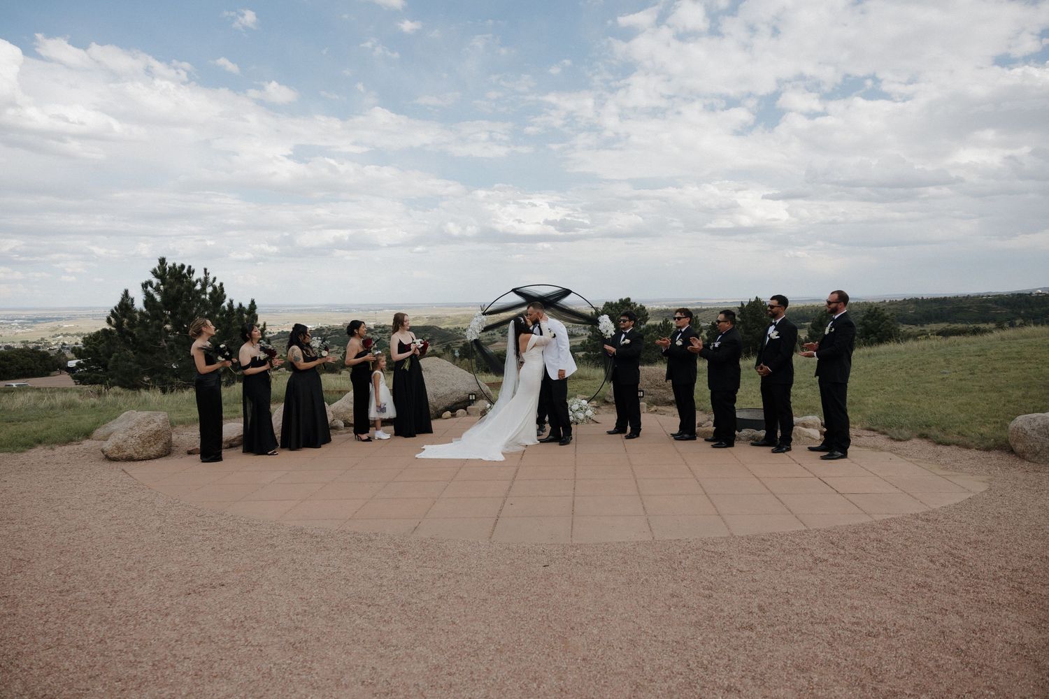 Wedding party in black attire stands with bride and groom under umbrella at scenic outdoor ceremony venue.