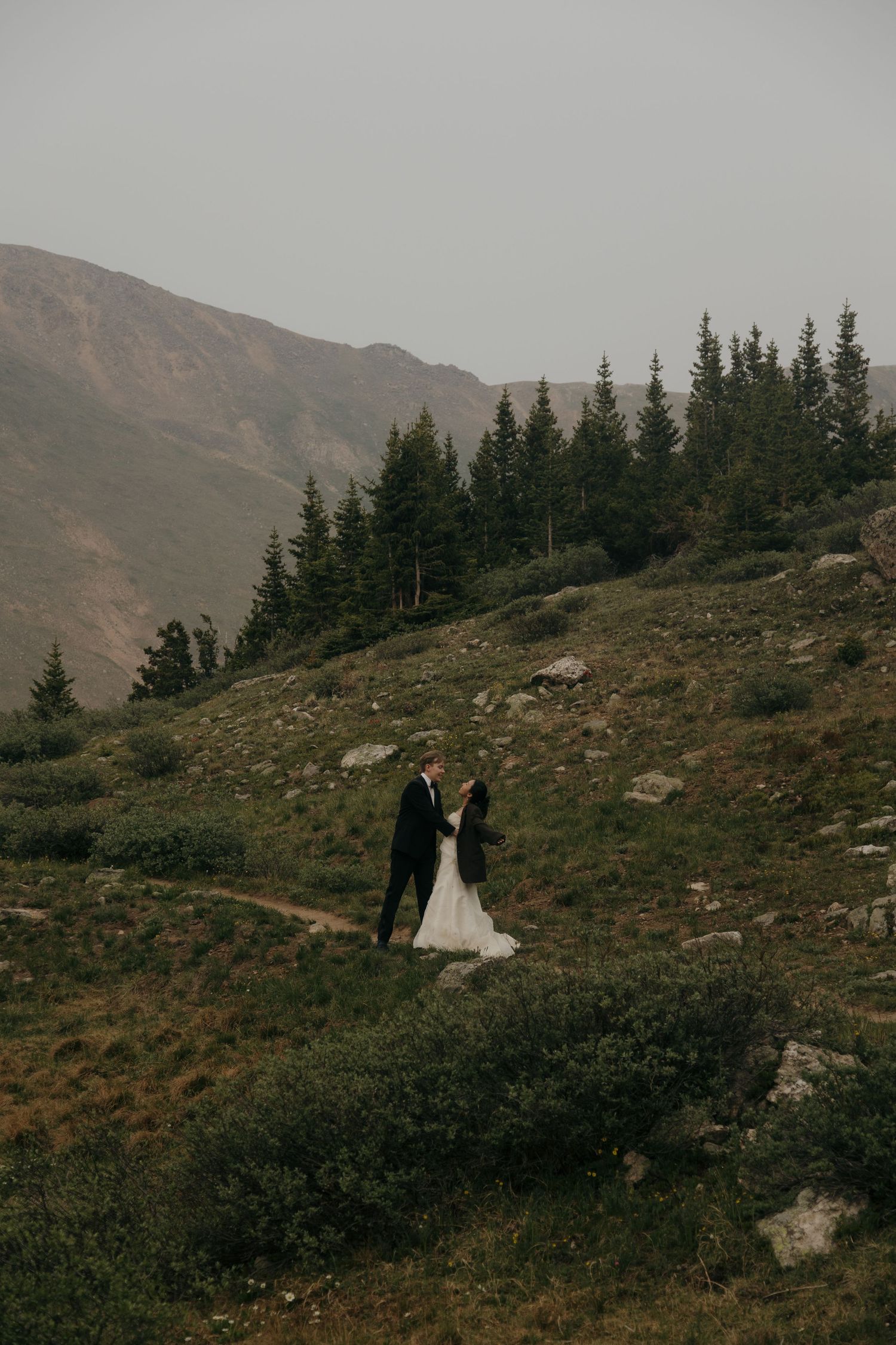 Bride and groom hold hands on a lush green mountain hillside surrounded by evergreen trees and rocks.