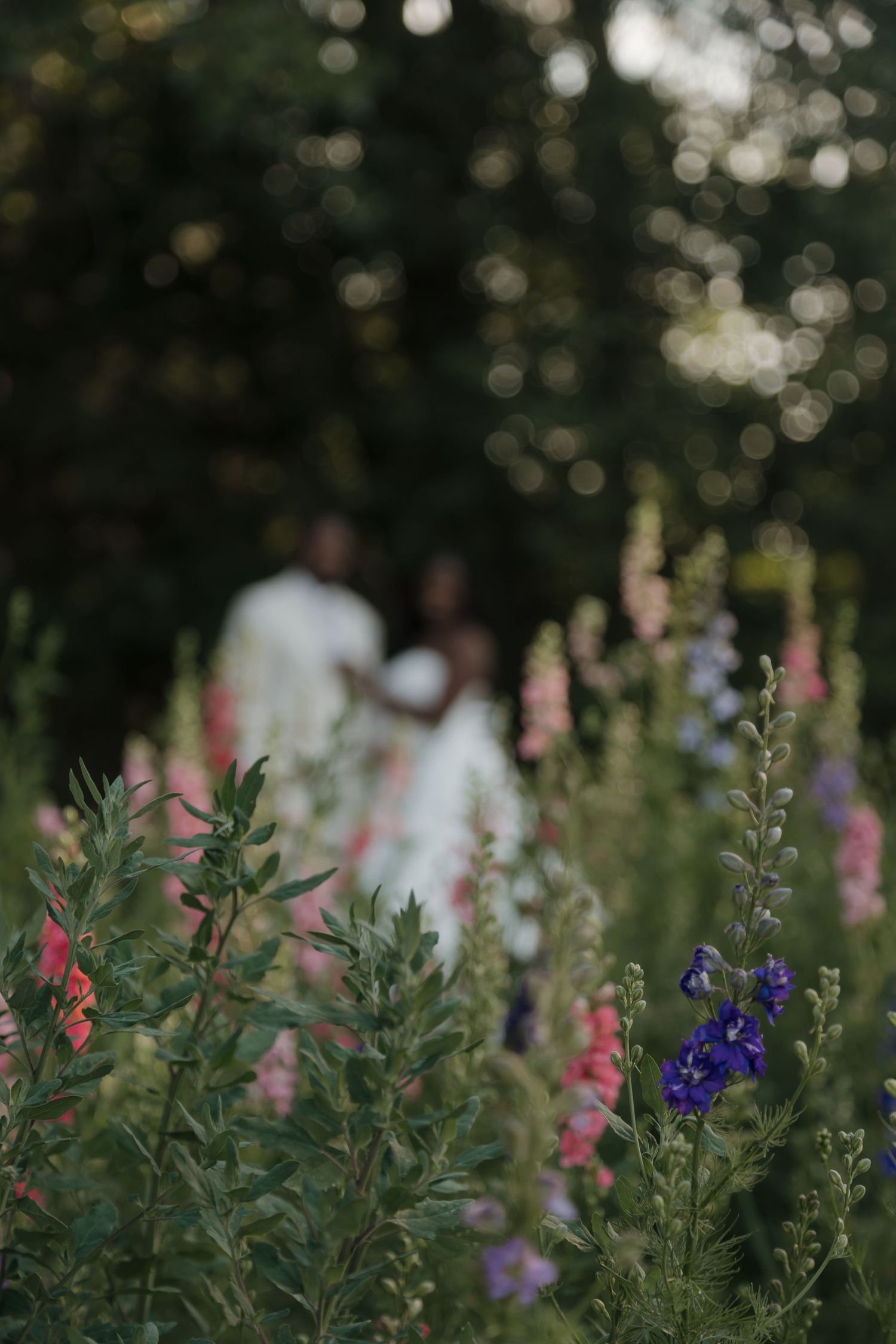 A couple stands blurred behind vibrant wildflowers in pink and purple at a garden wedding.