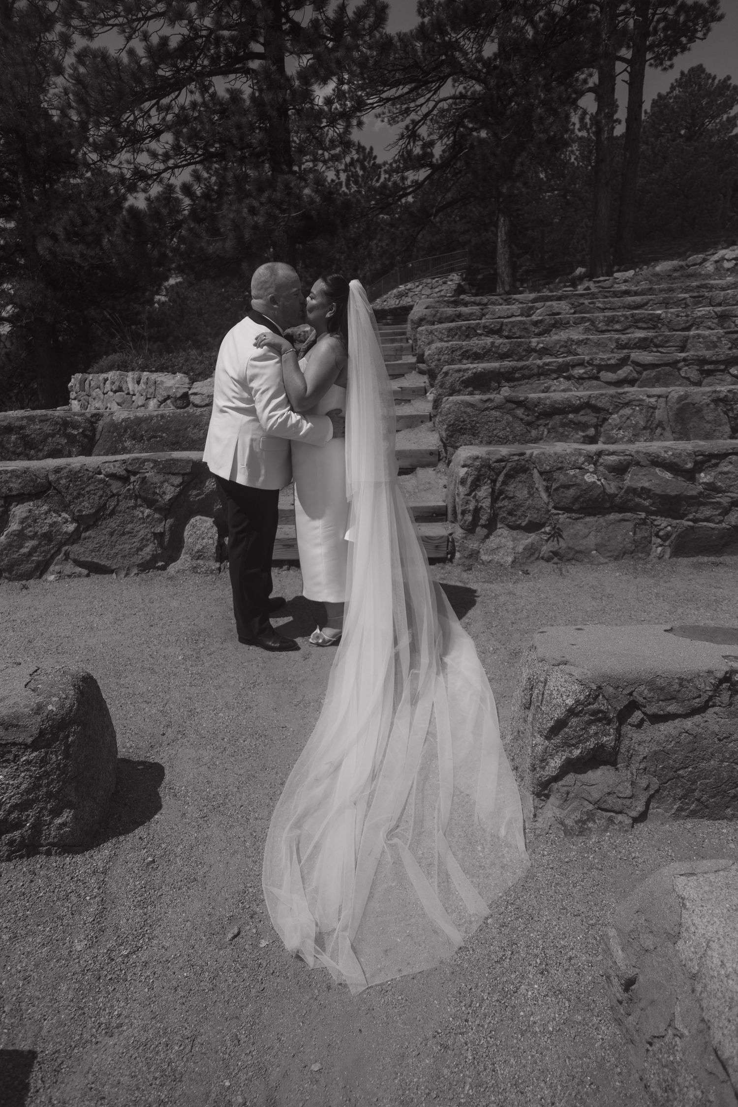 Black and white photo of a bride and groom embracing near stone steps, her long cathedral veil trailing on the ground.