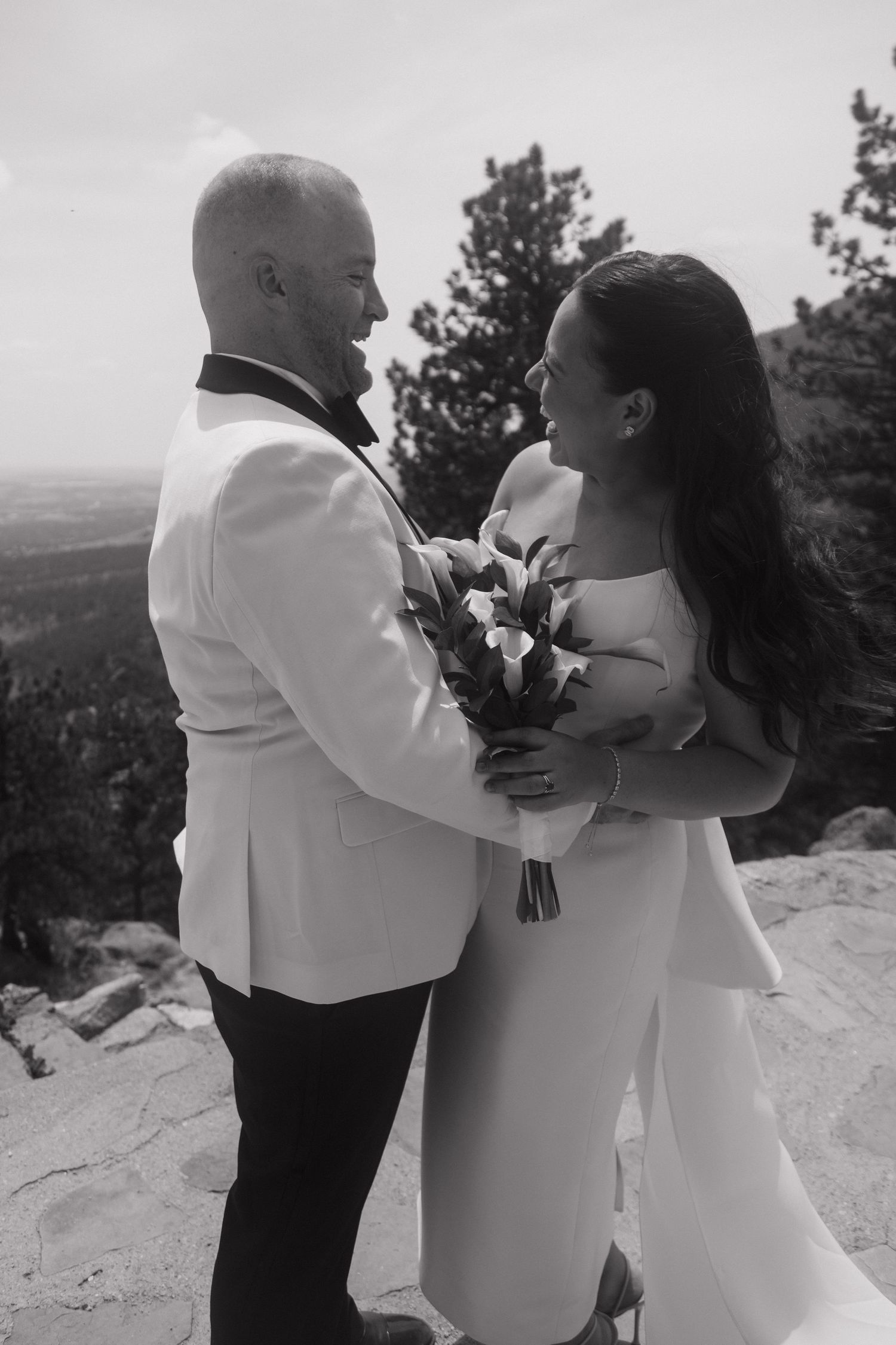 A black and white photo of a bride and groom sharing a joyful moment on a rocky mountain overlook.
