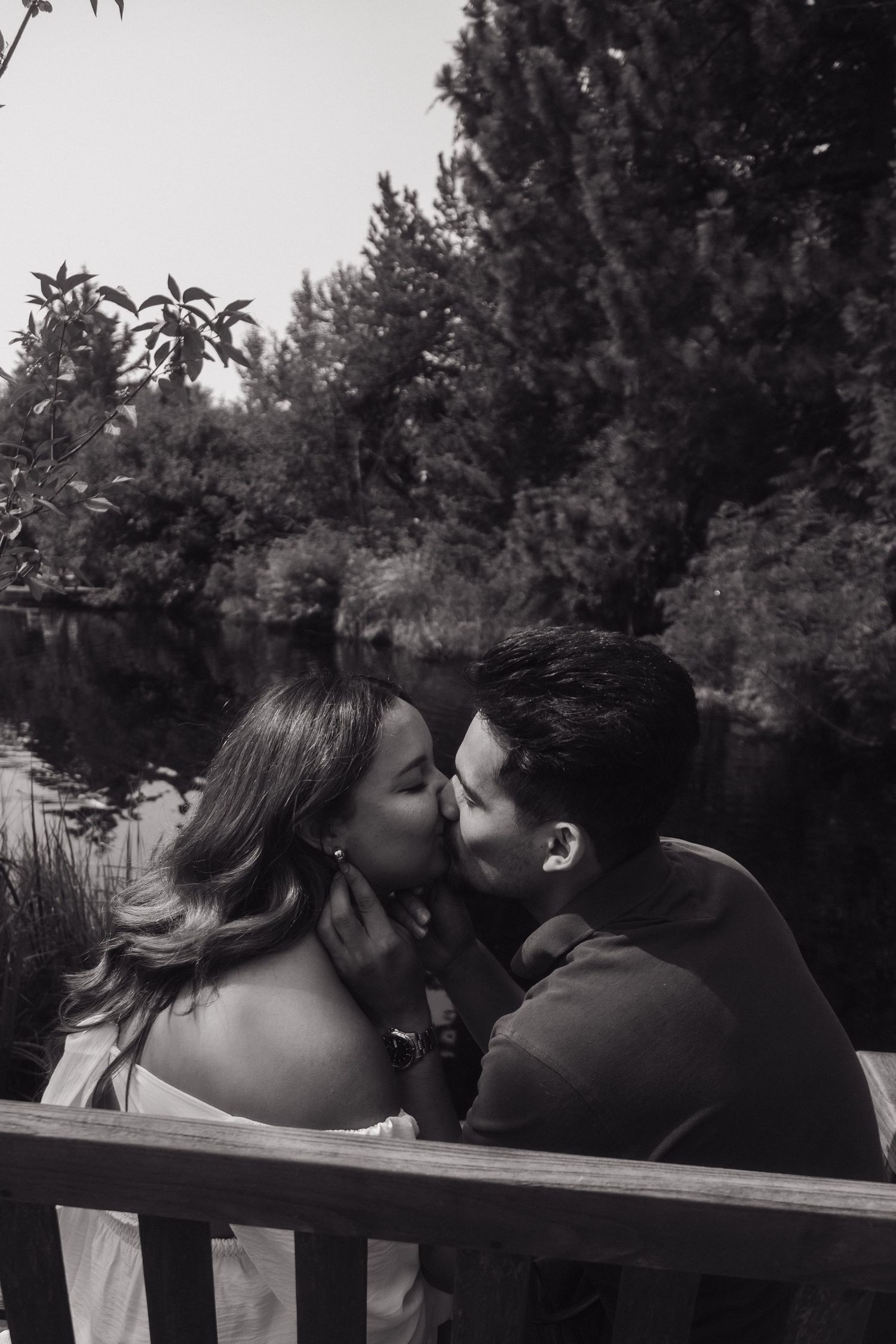 Black and white photo of a couple sharing a romantic kiss on a bench near a peaceful pond surrounded by trees.