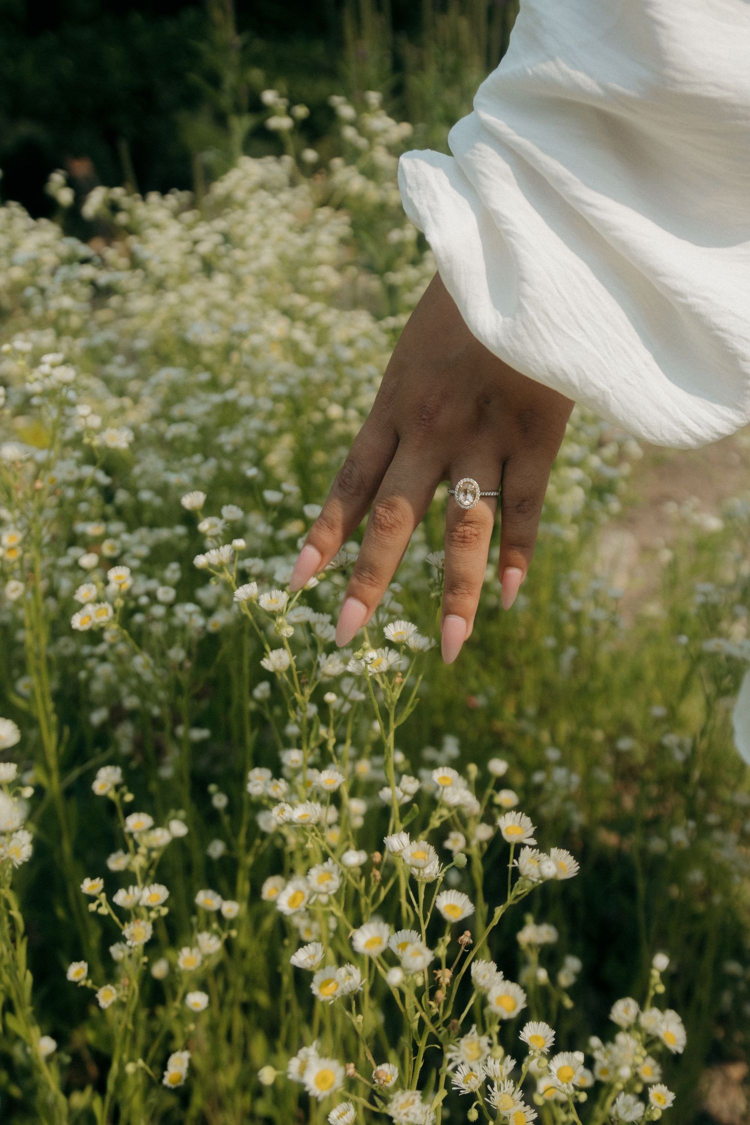 Close-up of a hand wearing an oval diamond engagement ring gently touching white wildflowers in a garden.