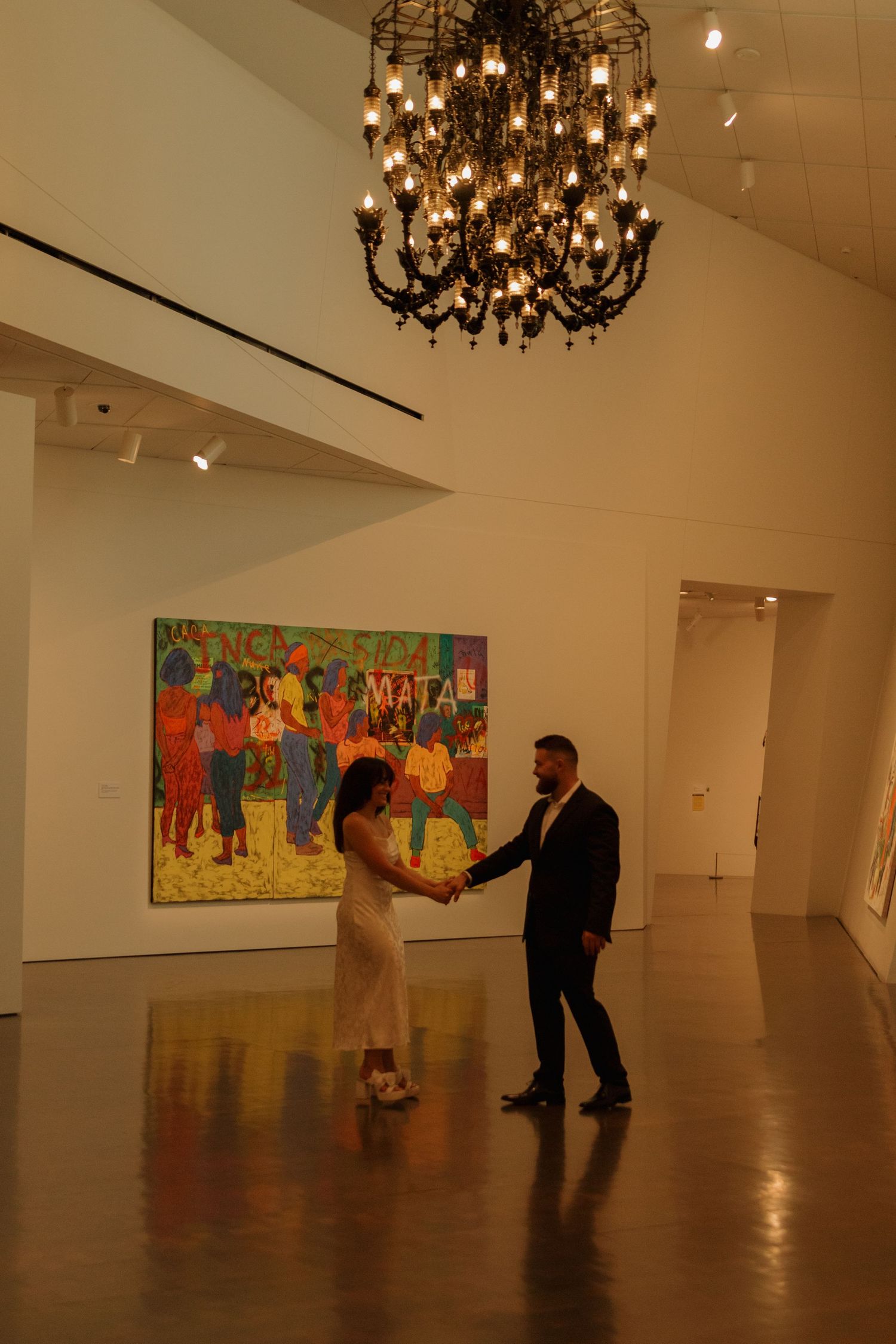 Two people dance in a spacious gallery with chandeliers and colorful artwork on display.