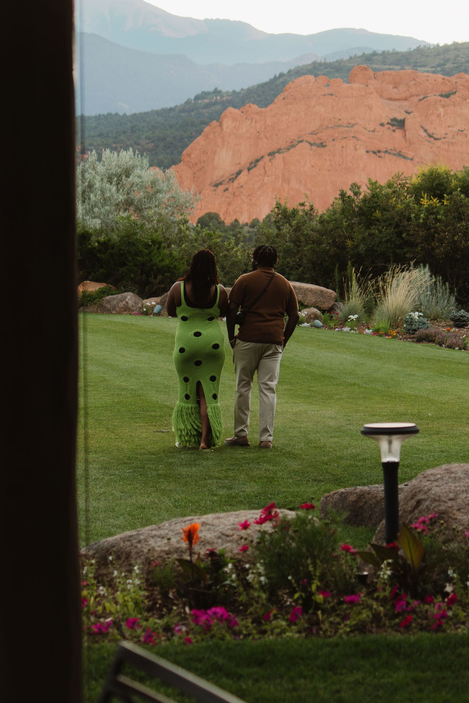 Couple walking on manicured golf course lawn with vibrant flower garden and red rock formations in background.